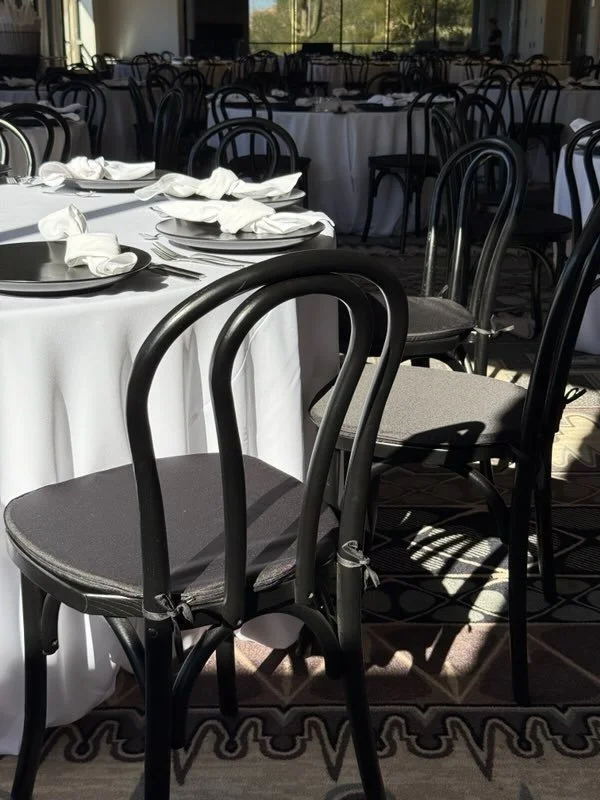 Empty banquet hall with round tables covered in white tablecloths, set with white napkins and plates, with black chairs arranged around the tables, sunlight shining through windows.