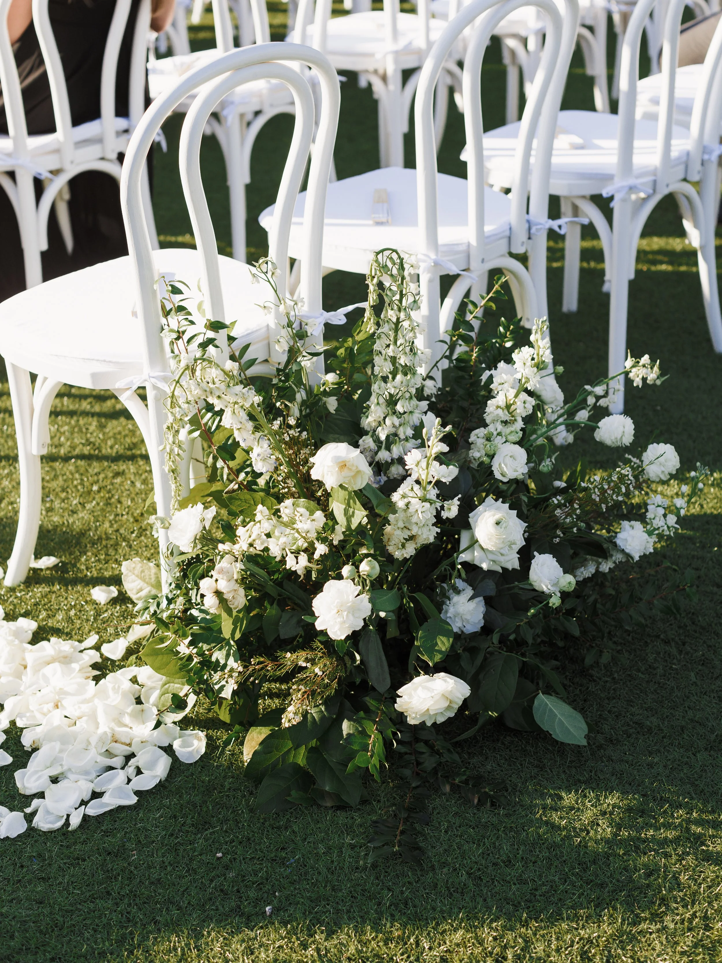 Outdoor wedding ceremony with white bentwood chair and white tie cushion setup with white chairs and a large white floral arrangement on green grass.