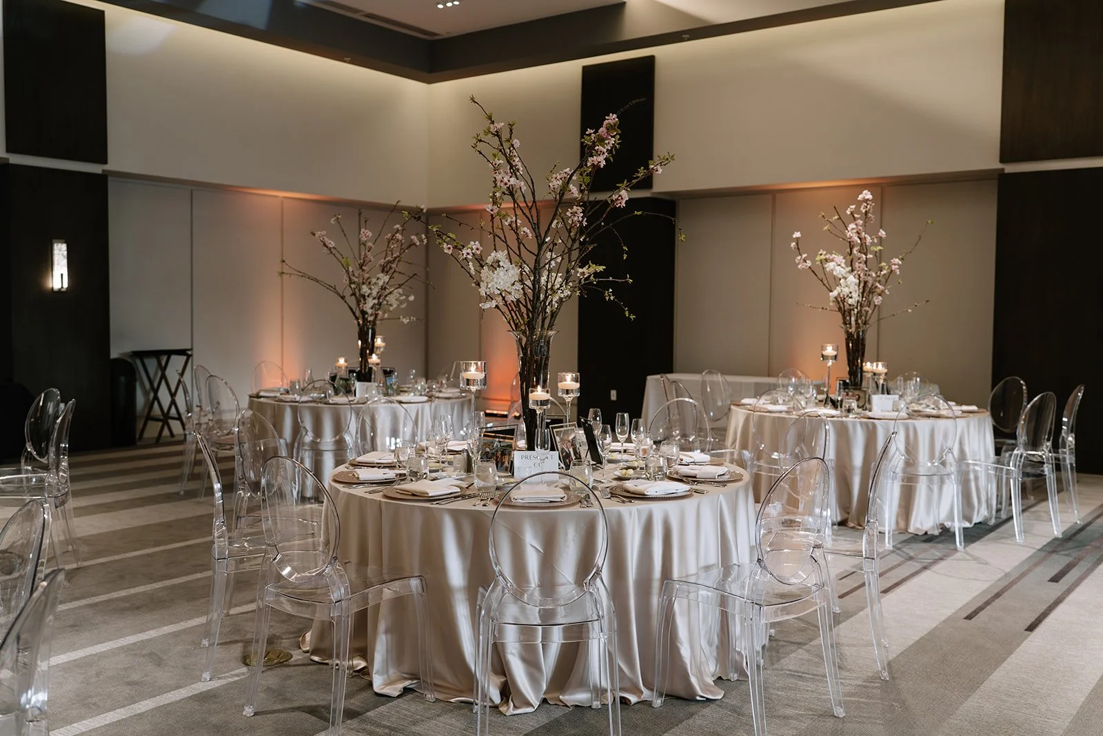 Elegant banquet hall with round tables covered in white tablecloths, decorated with large floral centerpieces of cherry blossoms and candles, set with ghost chairs for a formal event.