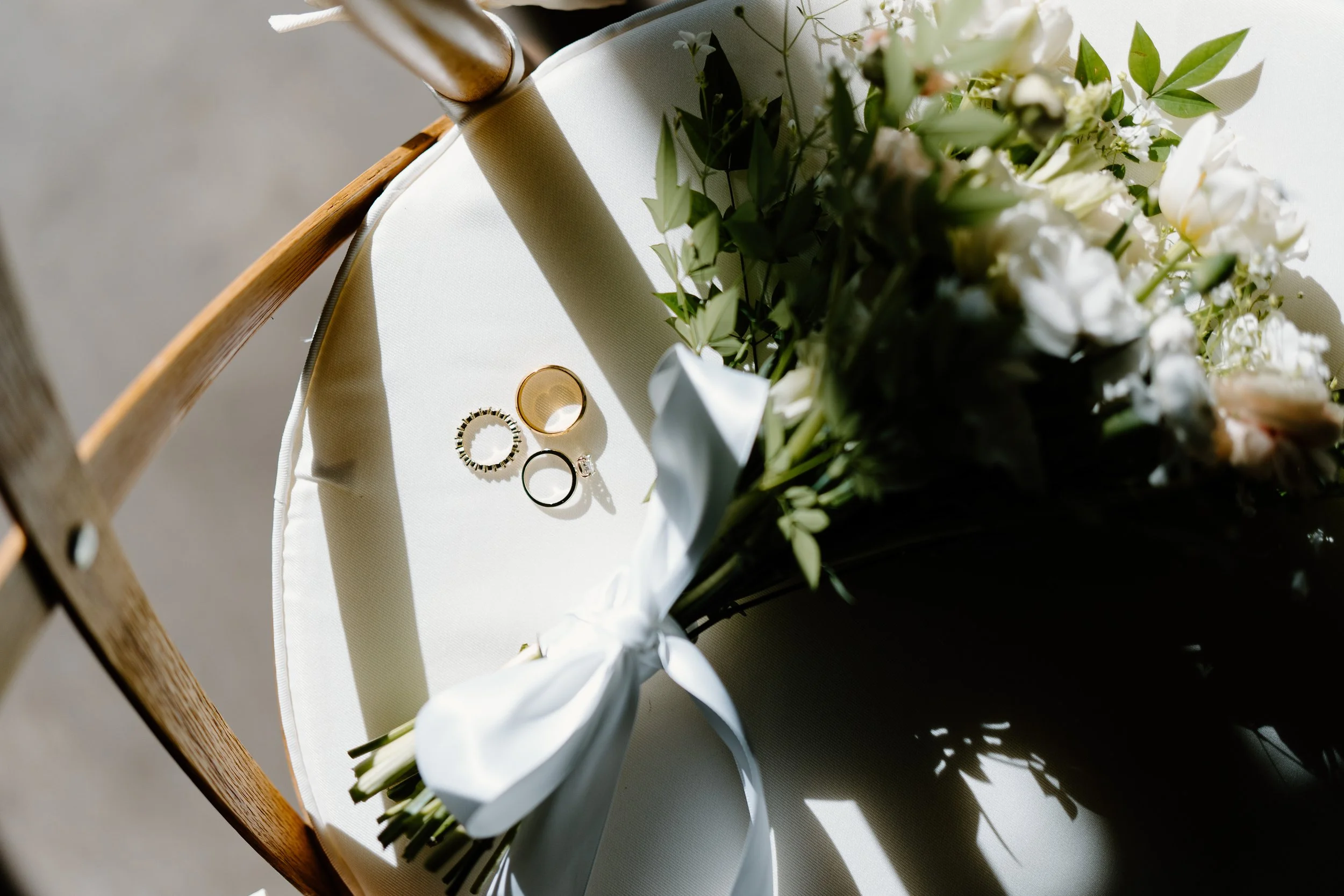 Close-up of wedding rings and a bouquet of white flowers on a natural wood x back chair, with sunlight casting shadows.