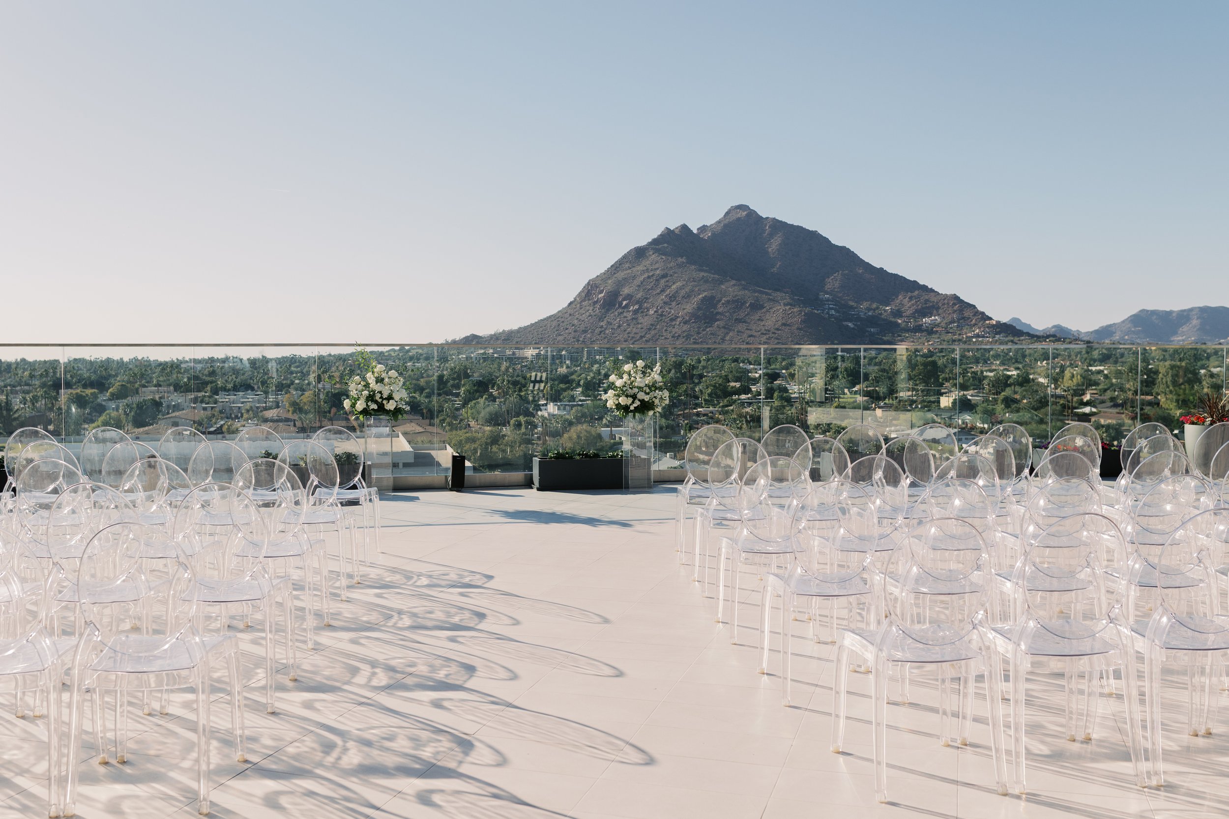 Outdoor event space with clear acrylic chairs arranged in rows, floral arrangements on stands, and a mountain in the background under a clear sky.