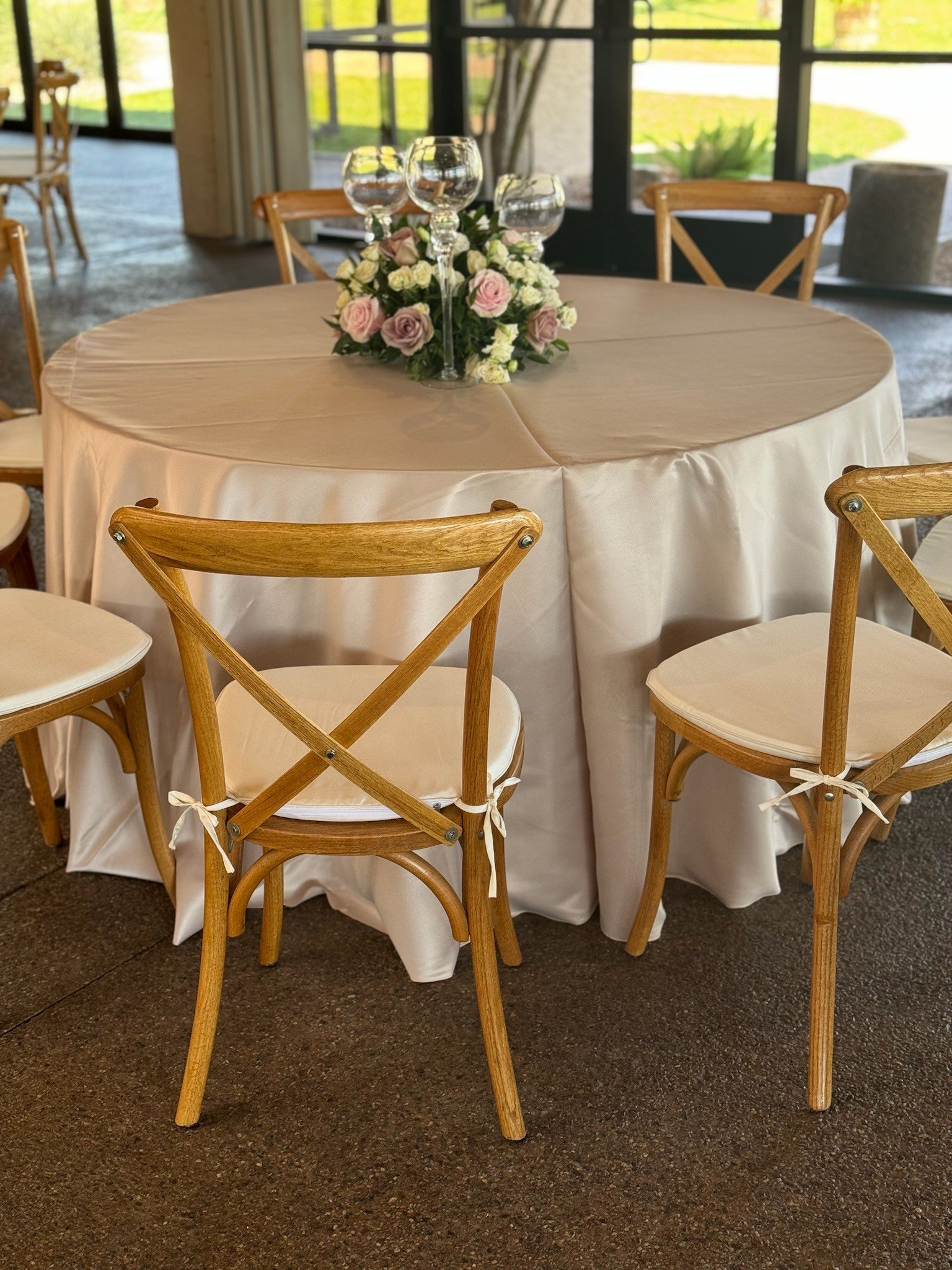 Round banquet table with a cream-colored tablecloth, with a floral centerpiece of pink and white roses, adorned with wine glasses. Xback / crossback chairs with cushioned seats in a bright, airy room with large windows and a view of a garden outside.