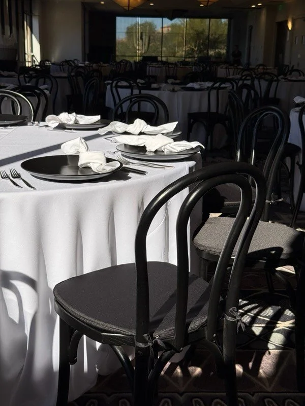 Wedding reception set with round tables set with white tablecloths, napkins, black plates, and silverware, with black bentwood chairs around the tables.