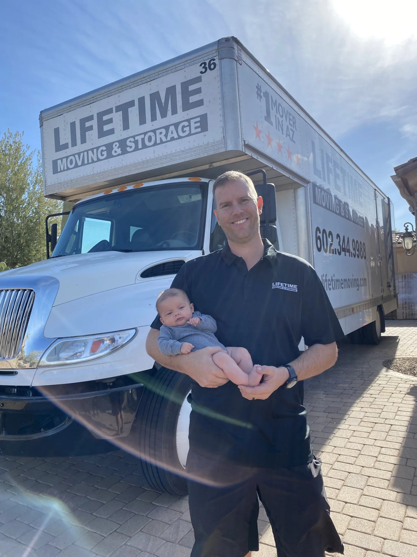 Man holding a baby in front of a moving and storage truck. The truck has "LIFETIME MOVING & STORAGE" written on it, along with contact information.