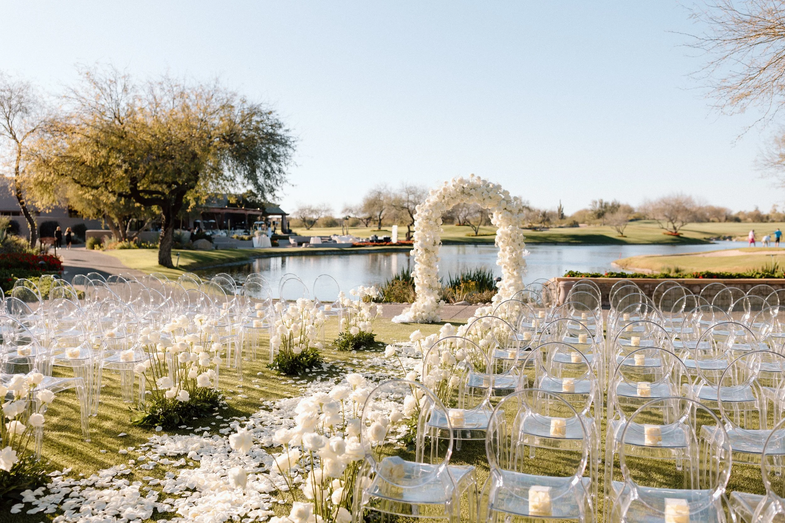 Outdoor wedding ceremony setup by a lake with transparent chairs, white floral decorations, and a floral arch, on a sunny day.