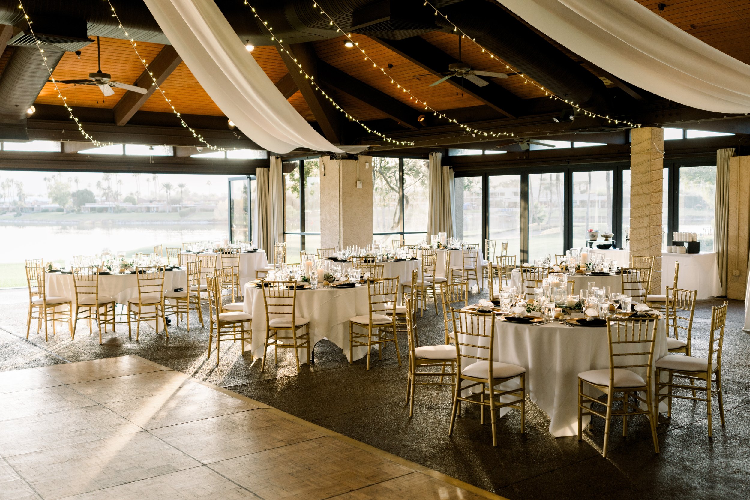 Elegant banquet hall with round tables set with white tablecloths, gold chairs, and glassware, decorated for an event with string lights and draped fabric on the ceiling. Large windows with a view of water and palm trees in the background.