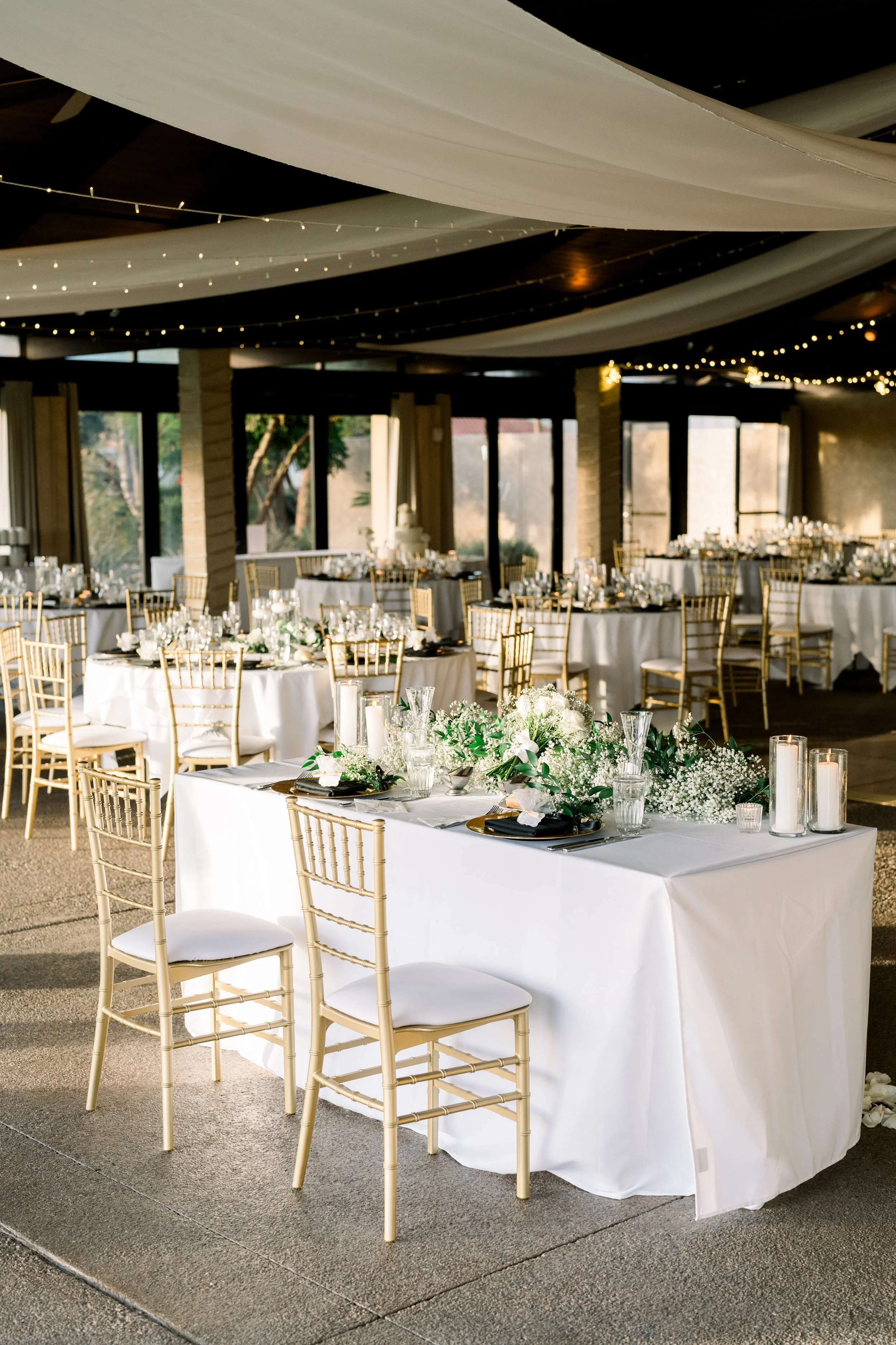 Elegant event space with decorated tables, gold chiavari chairs, floral centerpieces, and candles, prepared for a wedding or formal celebration.