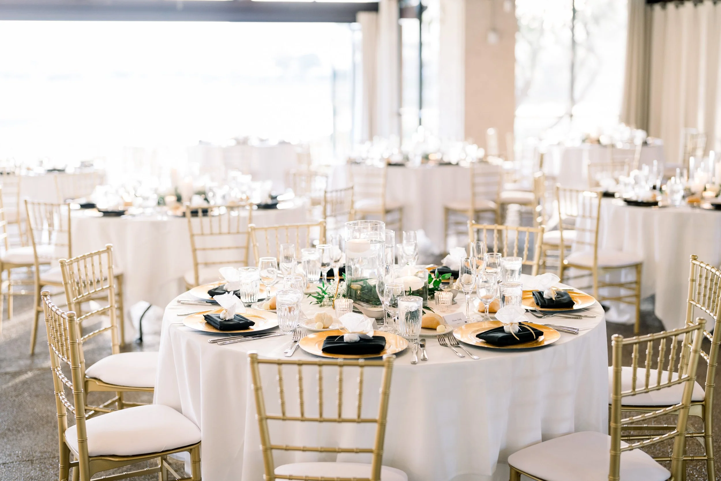 Elegant banquet hall with round tables covered in white tablecloths, set with gold charger plates, black napkins, glasses, and decorative centerpieces. Gold chiavari chairs the tables, and the room is brightly lit with natural light from windows.