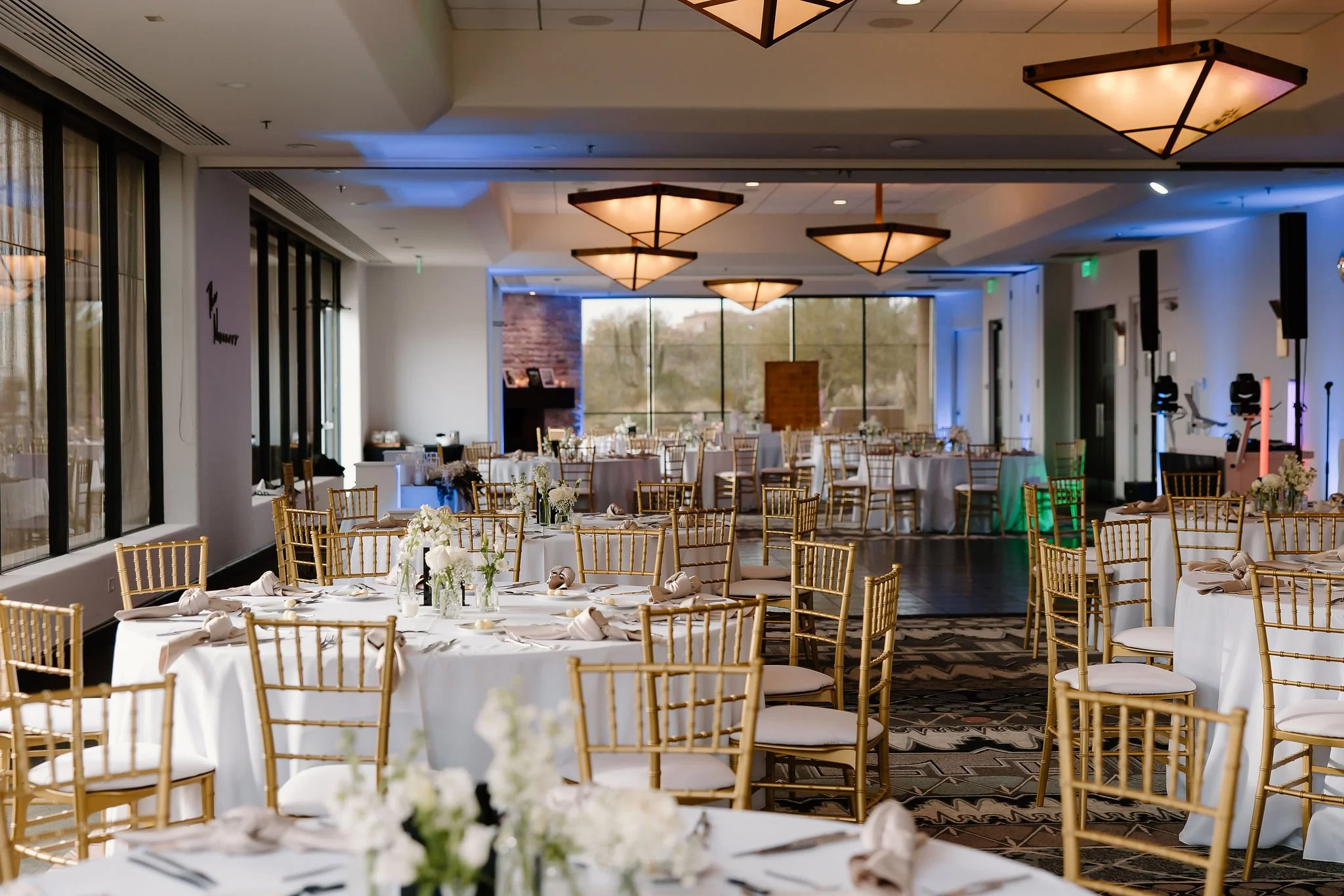 Elegant banquet hall decorated for a special event with round tables covered in white tablecloths, gold chiavari chairs, floral centerpieces, and large windows letting in natural light.