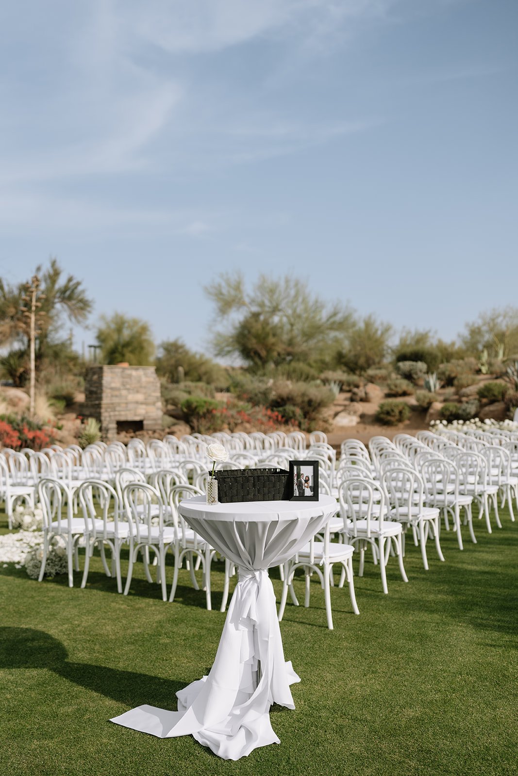 Outdoor wedding ceremony setup with white bentwood chairs arranged in rows, a small table with a white cloth, a framed photo, and a flower vase, set on a grassy area under a blue sky with sparse clouds.