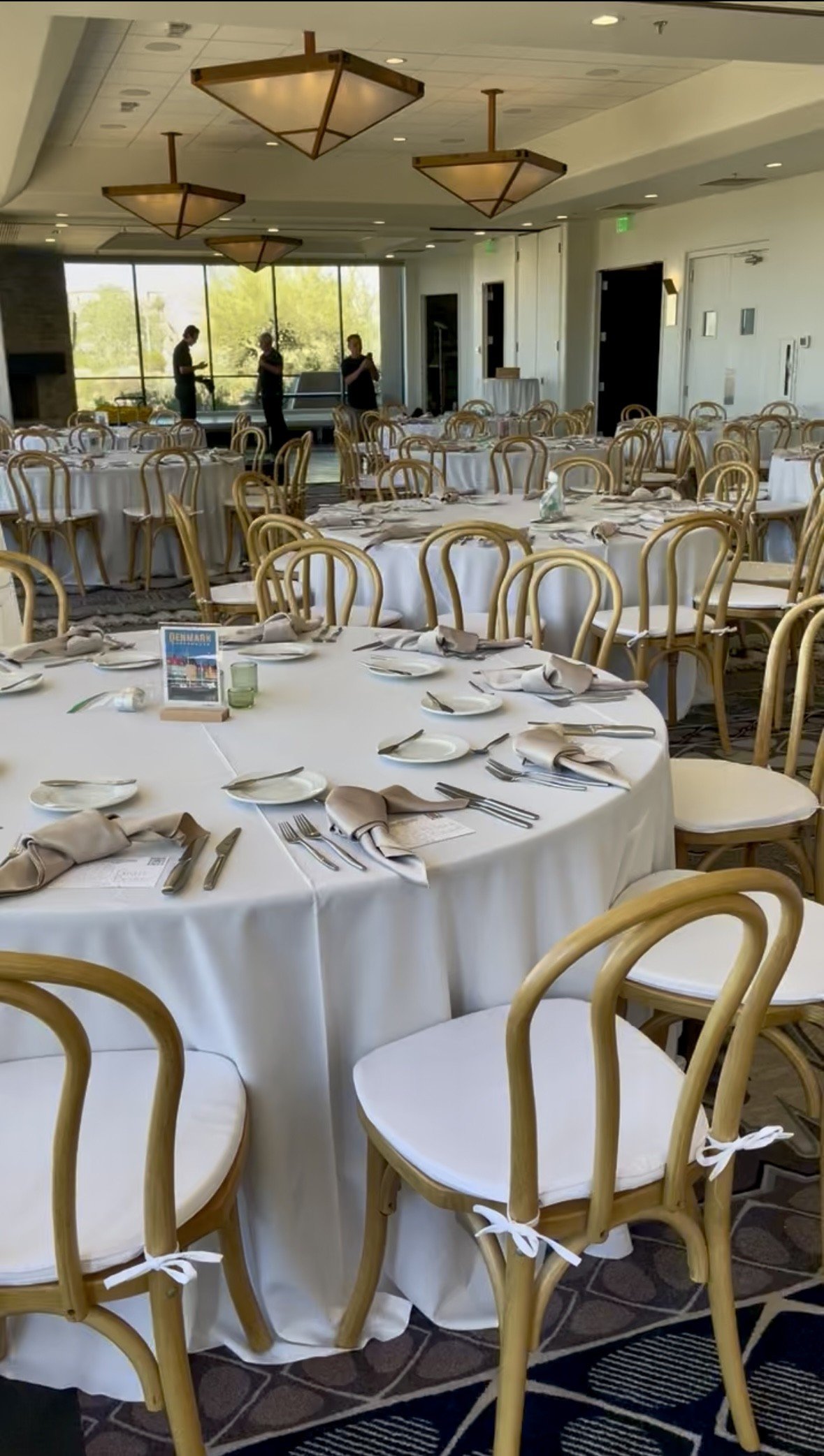 A spacious banquet hall with round tables set for an event, featuring white tablecloths, napkins, and cutlery, with natural wood bentwood chairs and white tie cushions. 