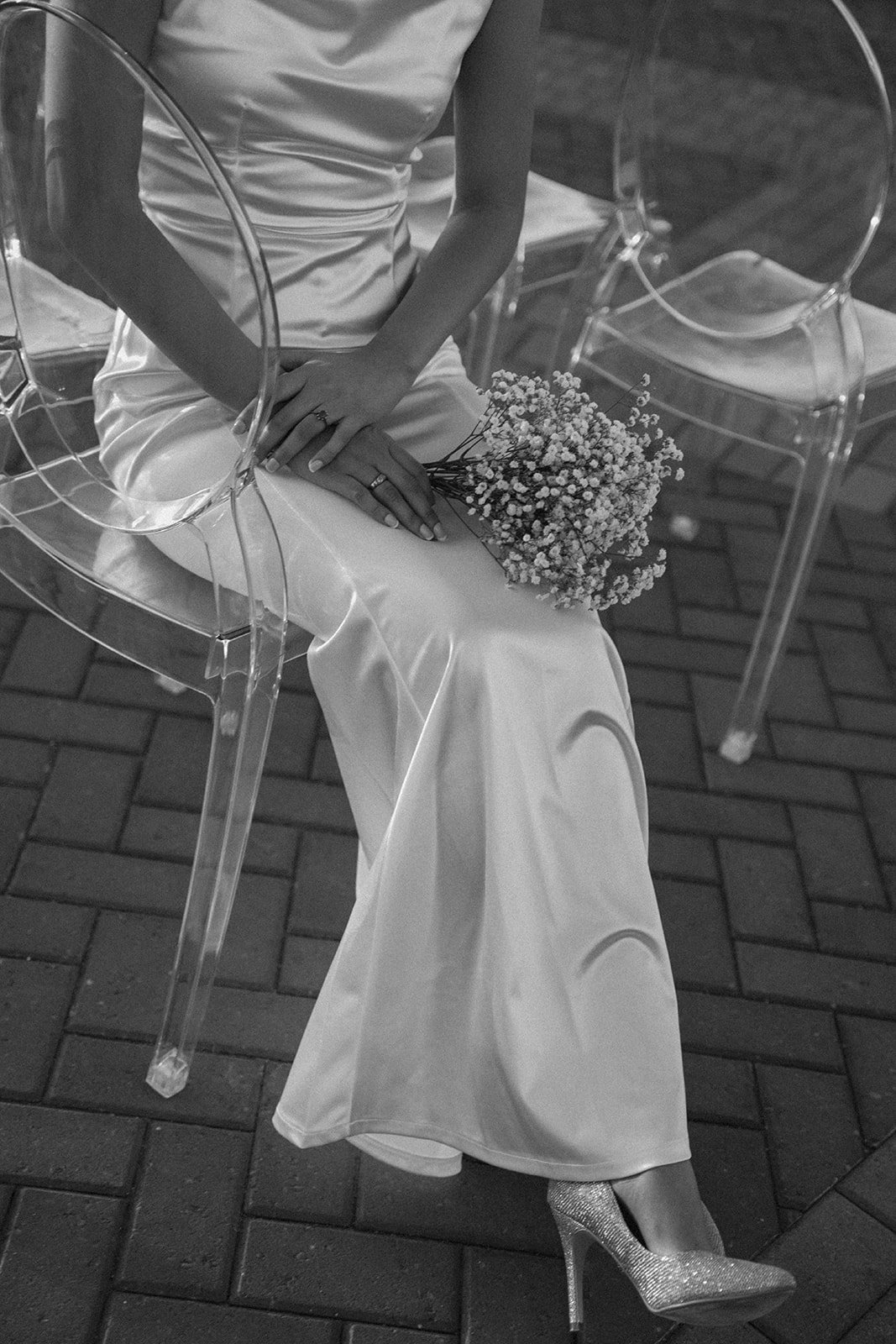 Woman in a satin dress sitting on a clear acrylic ghost chair, holding a bouquet of flowers, wearing glittery high heels, with another empty clear chair nearby, on a brick-paved surface.