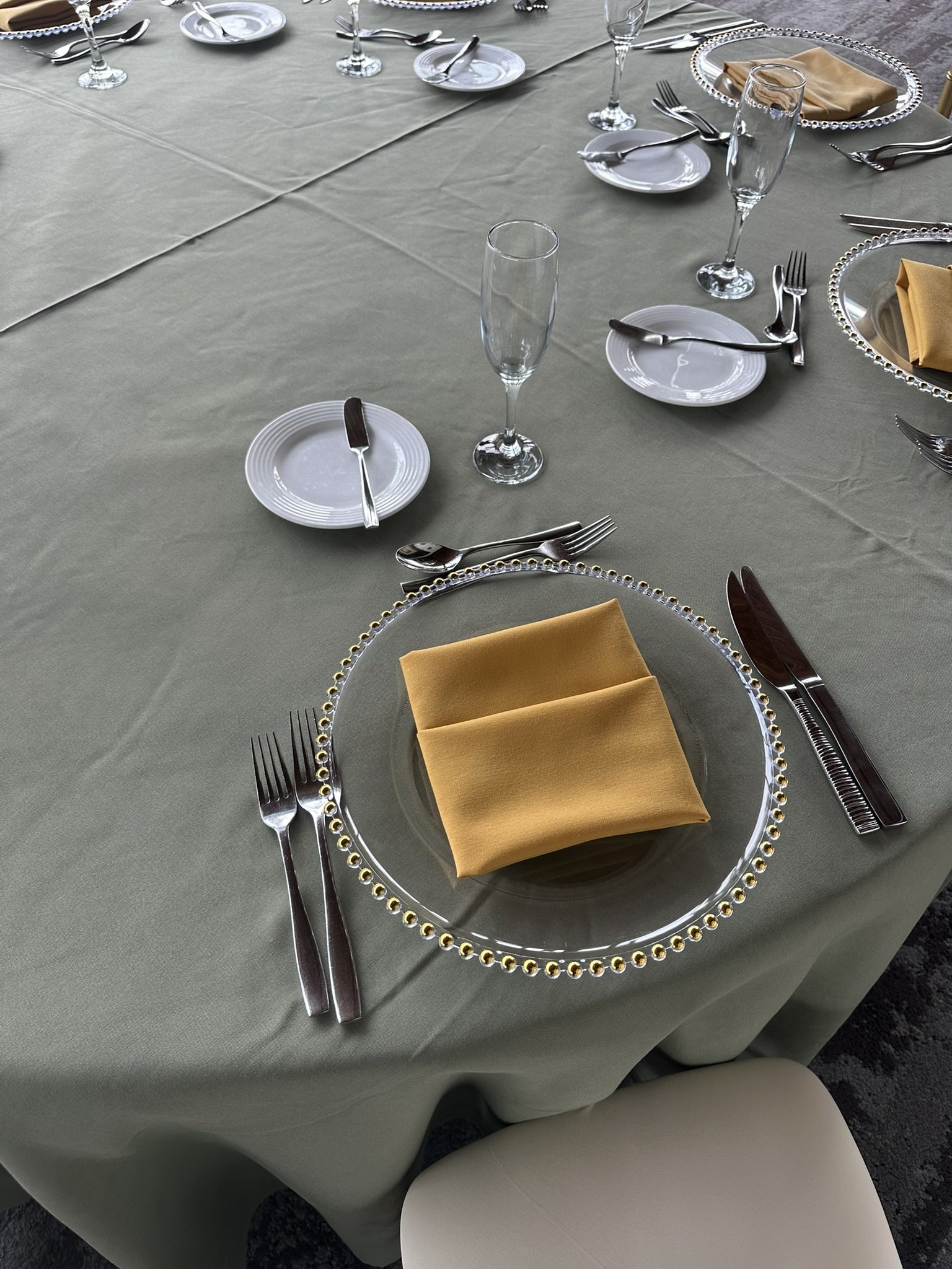 Formal dining table set with plates, napkins, silverware, clear charger plate with gold beads and glasses.