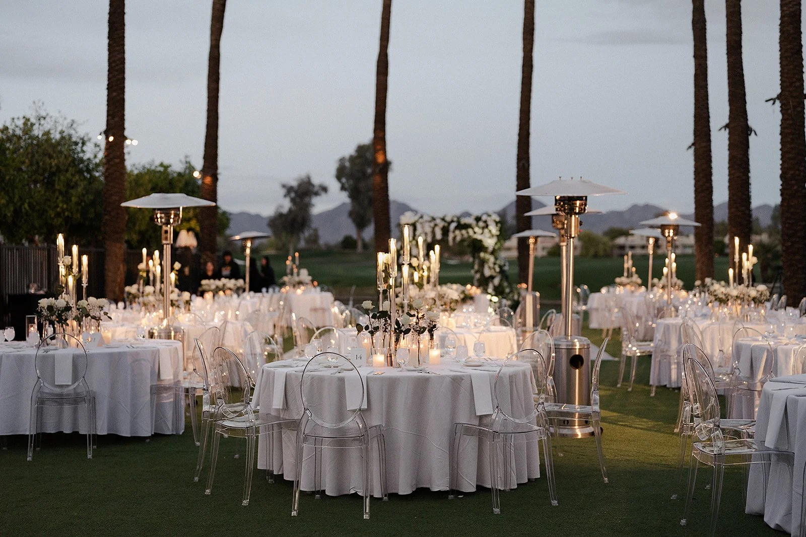 Elegant outdoor wedding reception setup with round tables draped in white tablecloths, surrounded by clear ghost chairs, elegant candlelit centerpieces, and tall patio heaters, with palm trees and mountains in the background during dusk.