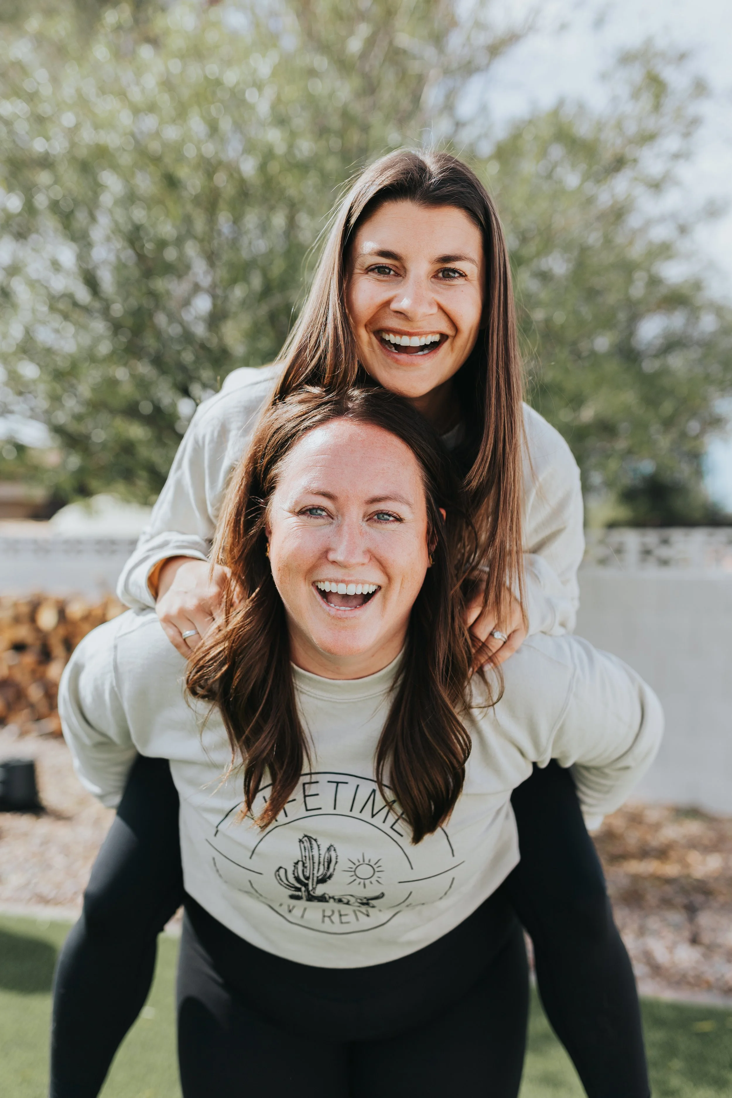 meet the owners and people behind the event rentals,  smiling, one on the other's back, outdoors with trees and a white fence in the background.