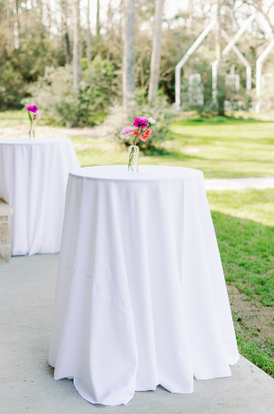 Outdoor setting with two high-top tables covered in white tablecloths, each decorated with a small flower arrangement, and a garden background with trees and a white fence.