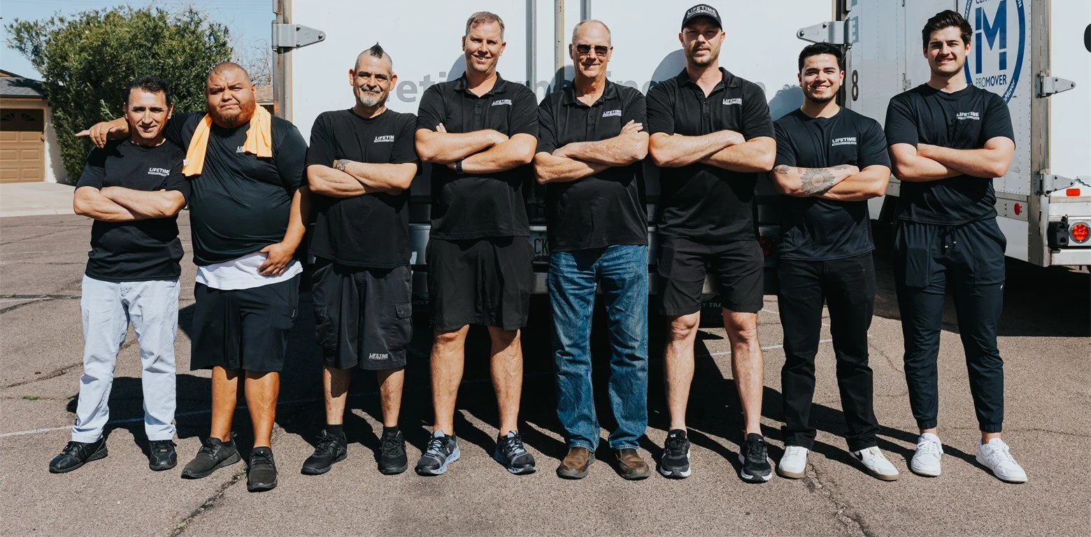 The Lifetime Moving and Event Rental Team - A group of eight men standing in a line outdoors, posing with arms crossed in front of truck trailers on a sunny day.