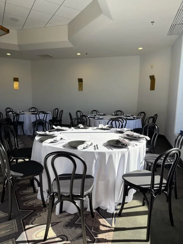 Ballroom with round tables covered in white tablecloths, black bentwood chairs, and place settings including plates, napkins, and silverware.