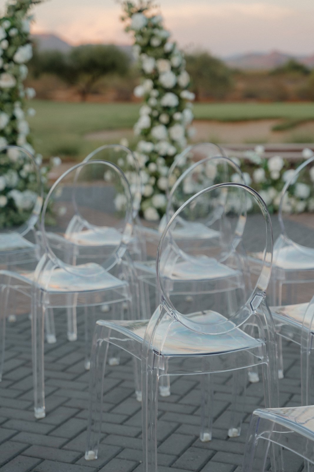 Clear acrylic ghost chairs arranged outdoors on a paved surface, with a floral arch and a blurred background of greenery and a sunset sky.