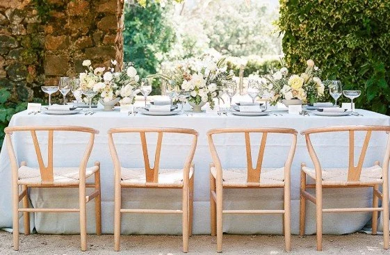 A long outdoor dining table set for a formal meal, with white tablecloth, multiple wine glasses, and floral centerpieces, surrounded by wishbone chairs, in a lush green garden.