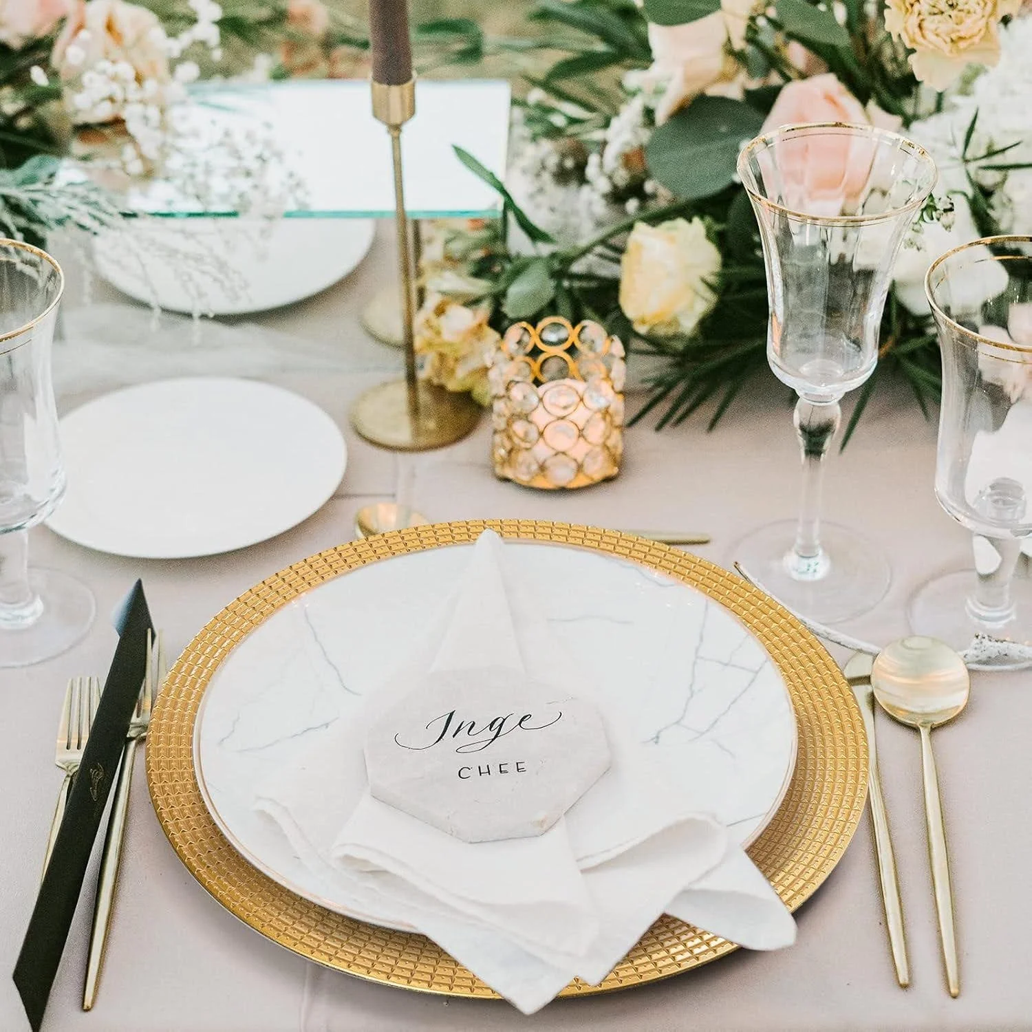 Elegant table setting with gold-rimmed glassware, a gold textured charger plate, a white napkin, and a personalized name card. The background features floral arrangements with roses and greenery, a decorative candle holder, and a table number stand.