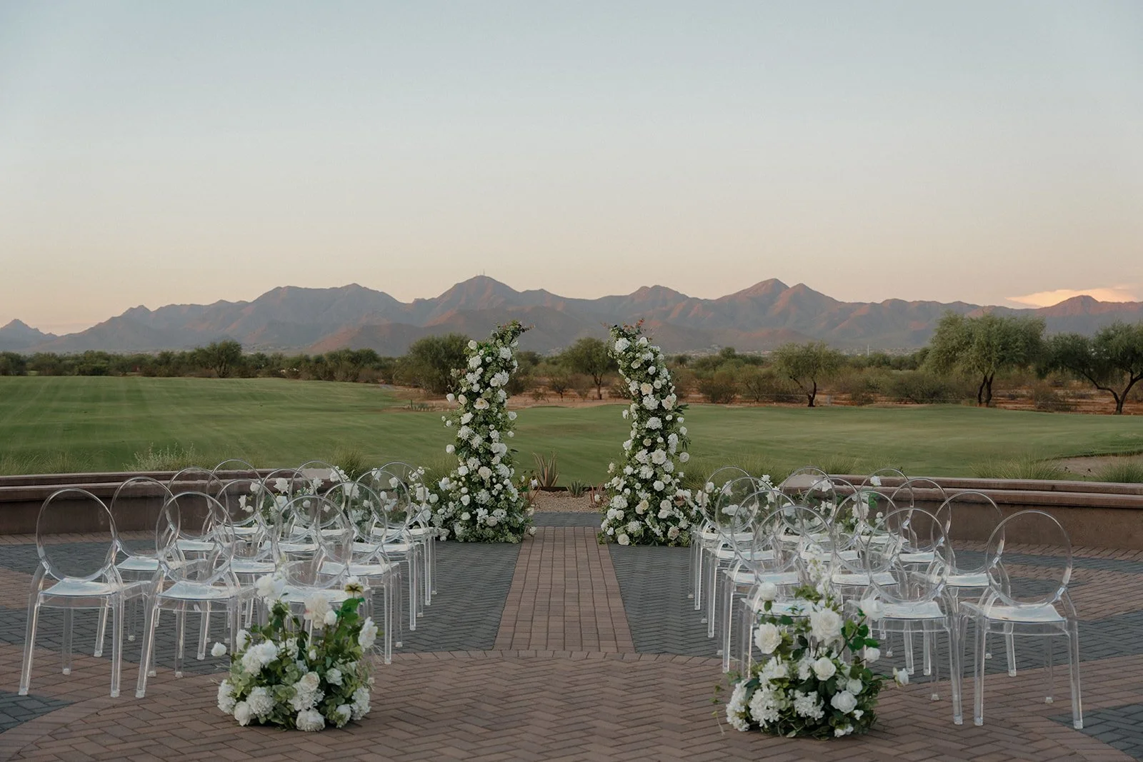 Outdoor wedding ceremony setup with ghost chairs white floral arches and chairs in a scenic desert landscape with mountains in the background. In Scottsdale, Arizona at the talking Stick Golf Club Resort 
