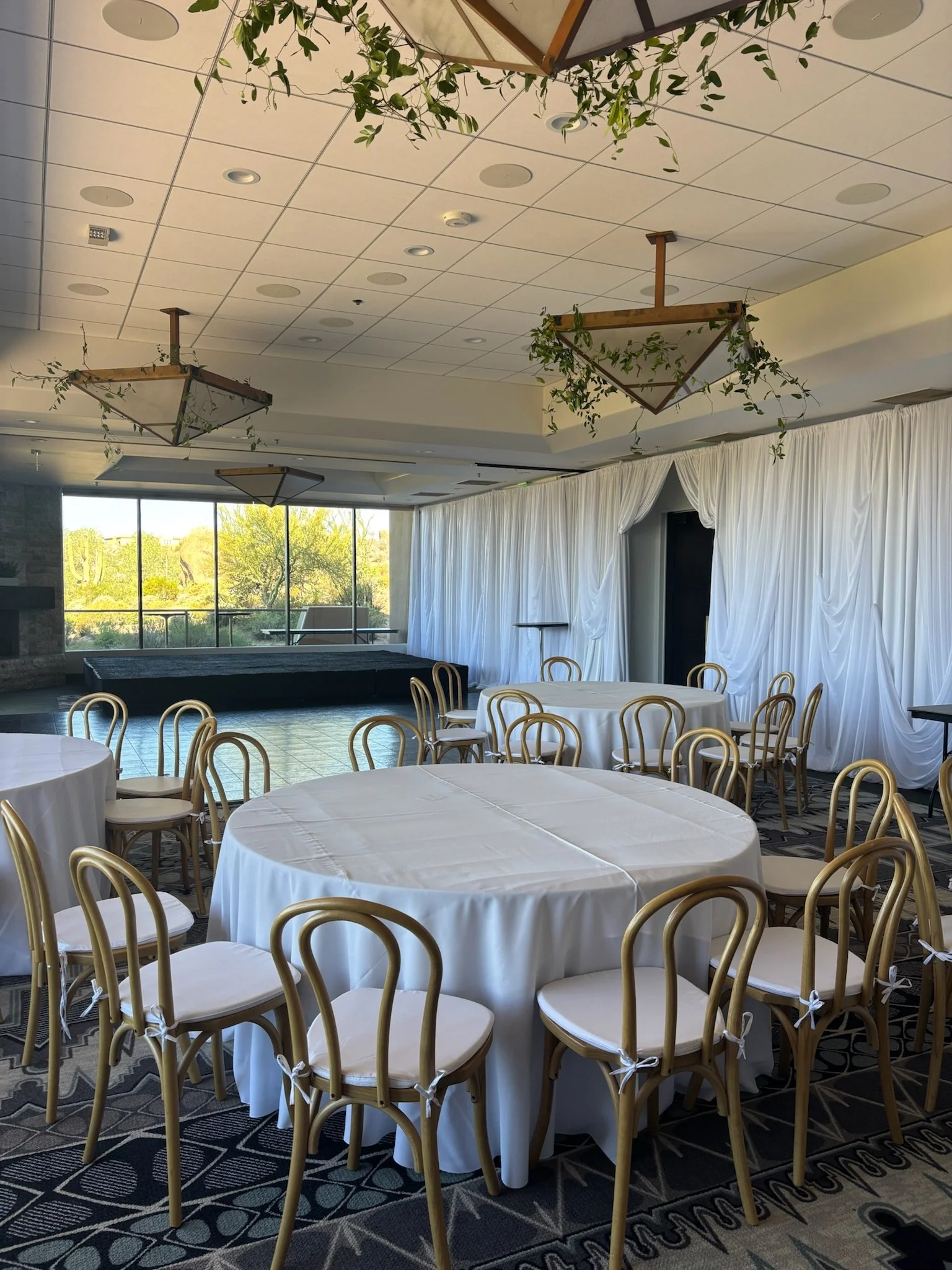 A decorated event space with round tables covered in white tablecloths, surrounded by natural bentwood chairs tied with white ribbons. Stage set white drapes hanging on the wall behind the stage. Greenery hang from the ceiling.