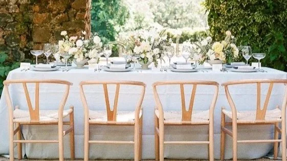Elegant outdoor dining table set with white tablecloth, glassware, and floral centerpieces, surrounded by wooden chairs with white cushions, on a patio with greenery.