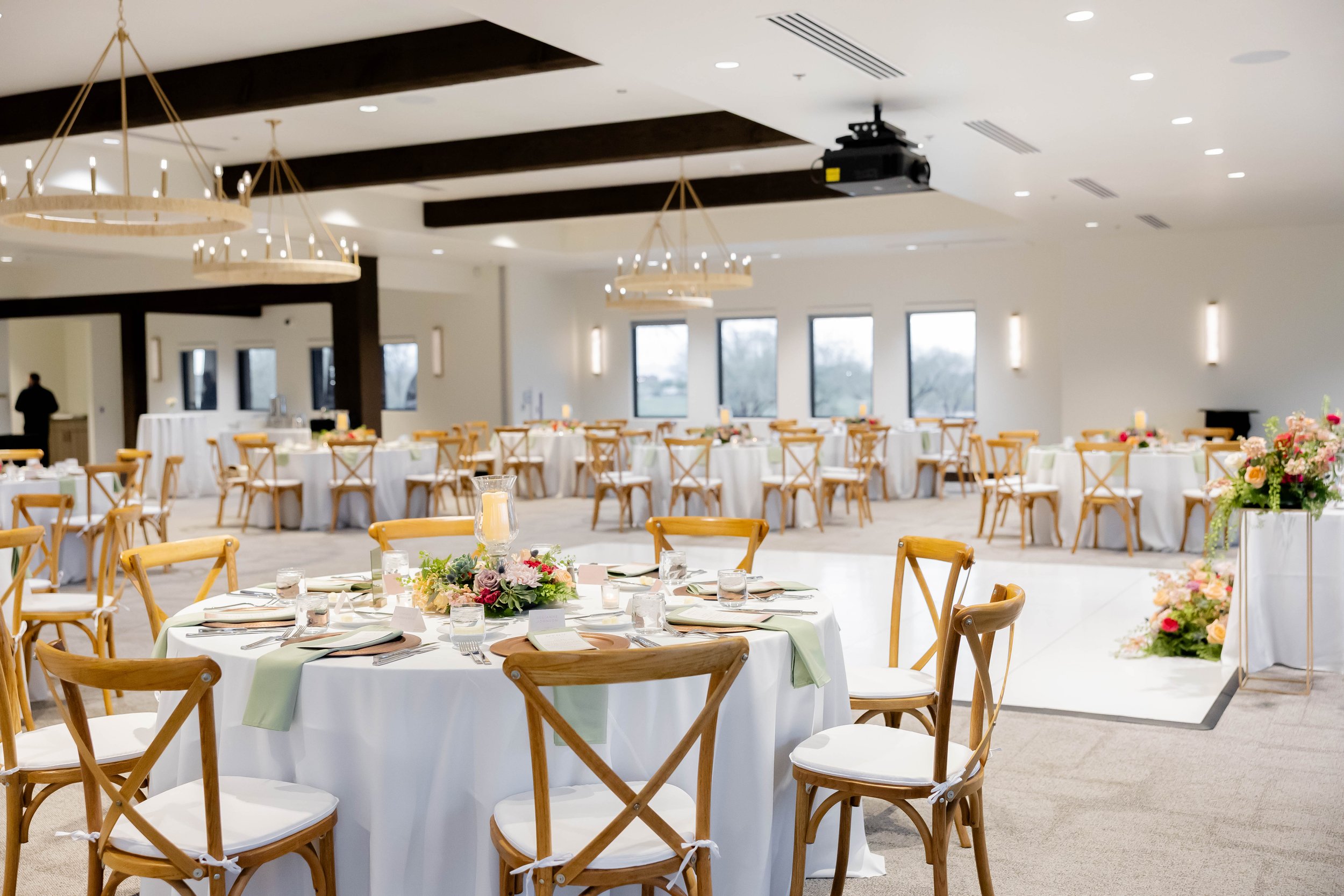 Event hall decorated with round tables with white tablecloths, floral centerpieces, and natural cross back wooden chairs, featuring large windows and modern lighting fixtures.