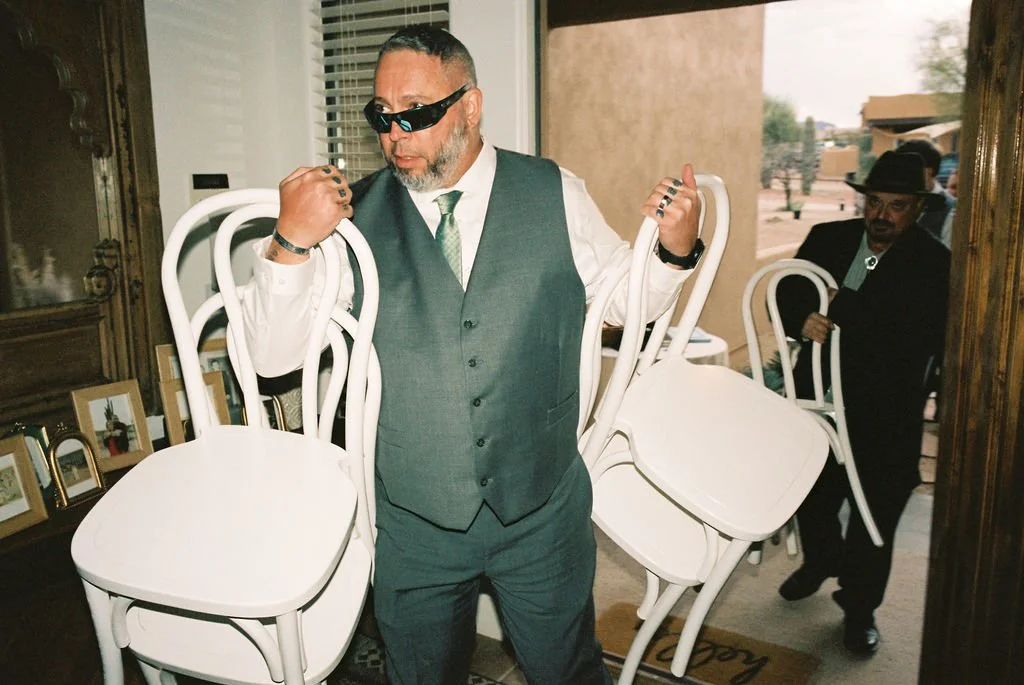 Two men dressed in suits and hats carrying white bentwood chairs, flipping ceremony chairs to intimate patio wedding reception.