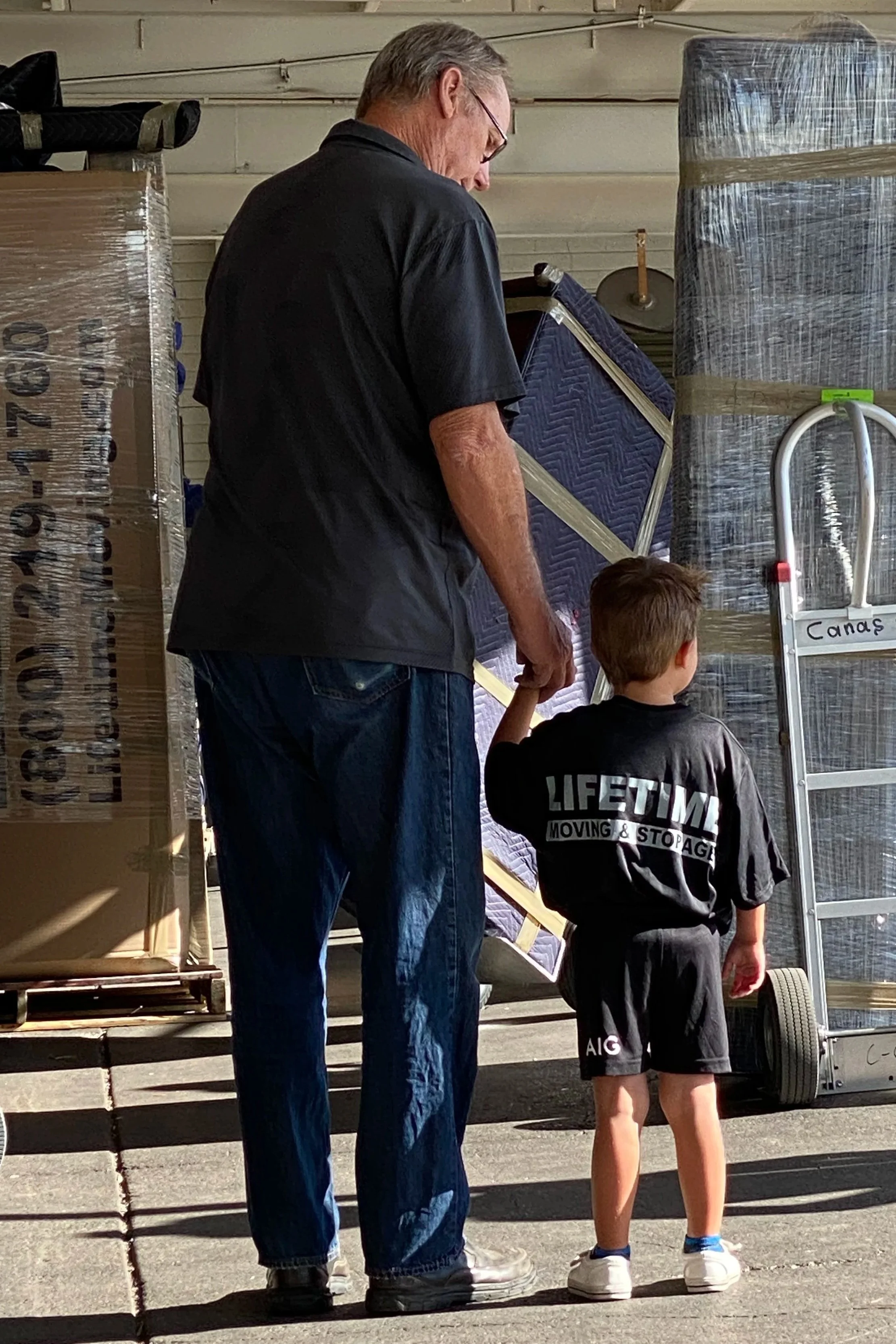 An older man and a young boy holding hands, with the boy wearing a black T-shirt and shorts, standing in front of moving boxes and a dolly, in a warehouse or storage area.