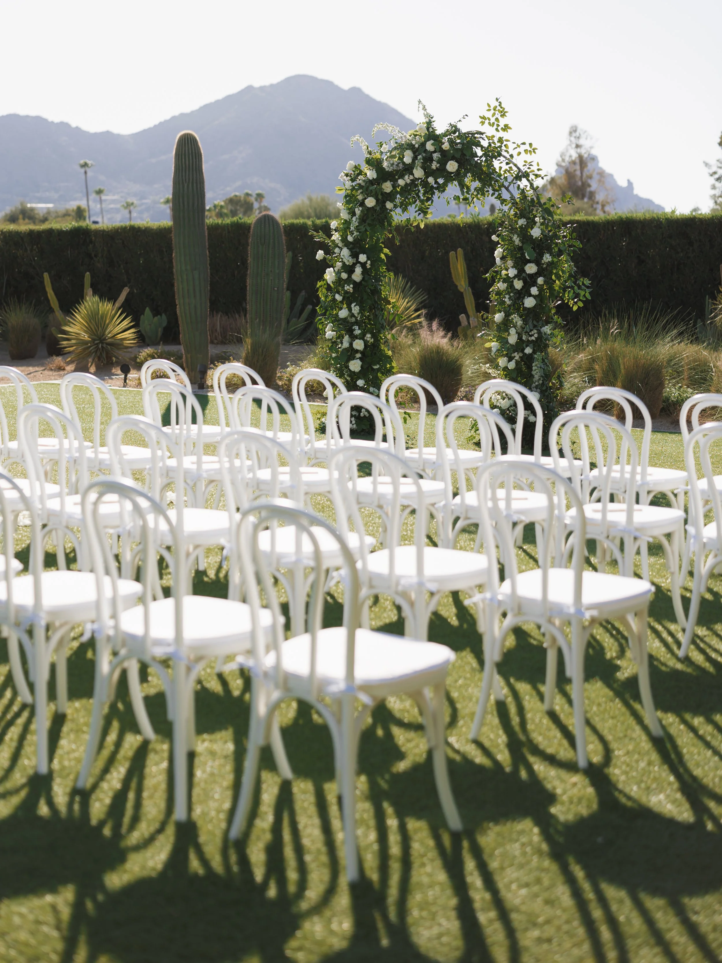 Outdoor wedding ceremony setup with white chairs arranged on grass, a floral arch, and desert plants and mountains in the background.