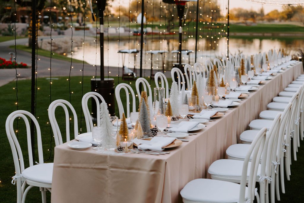 Long outdoor banquet table set with white plates, glasses, napkins, and festive decorations, set with white bentwood chairs and string bistro lights and water in the background during sunset.