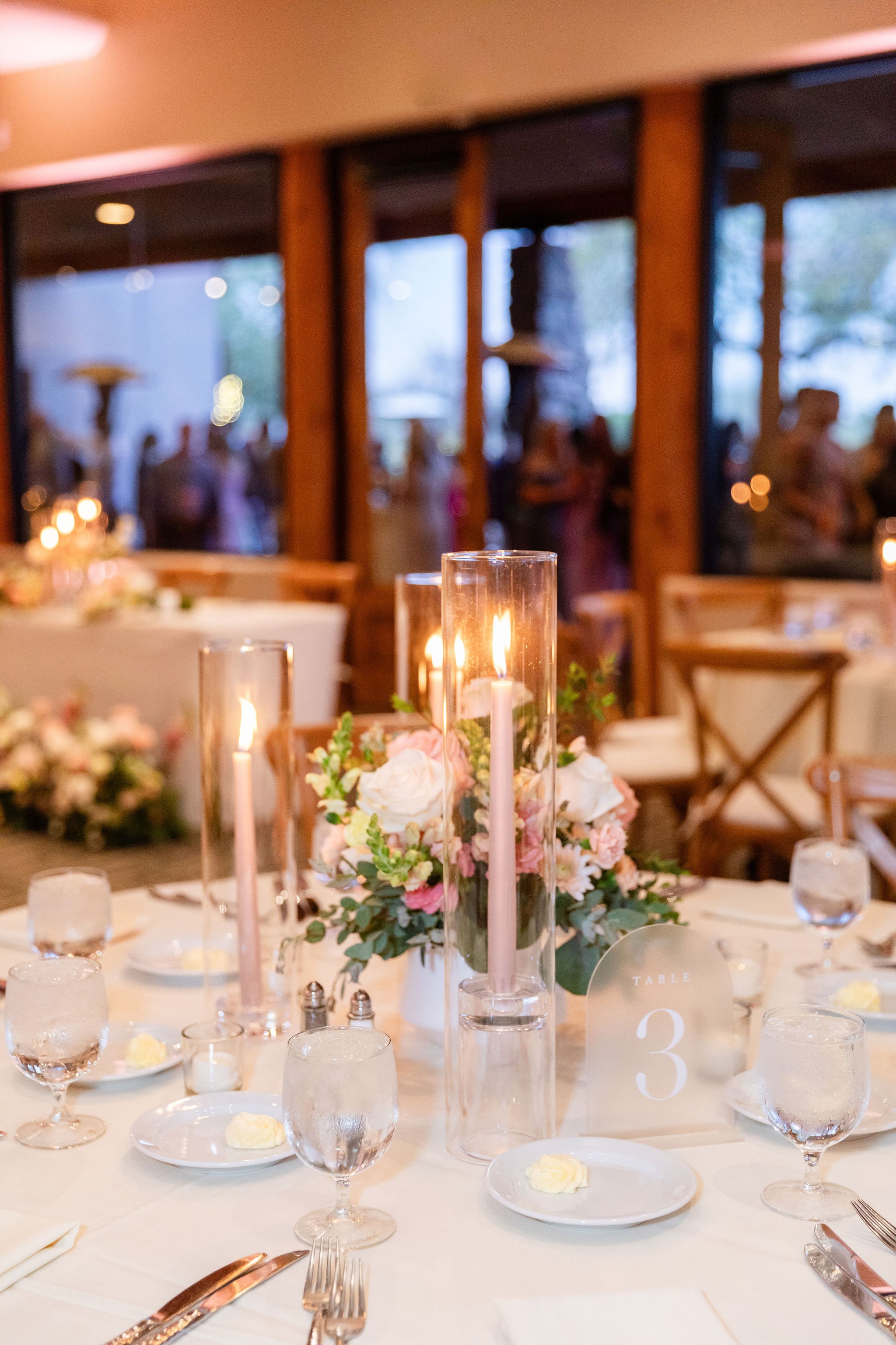 Elegant wedding reception table decorated with pink and white flowers, tall hurricane glass candleholders with pink candles, water glasses, plates with butter, and cutlery, with a large window behind showing guests and outdoor scenery.