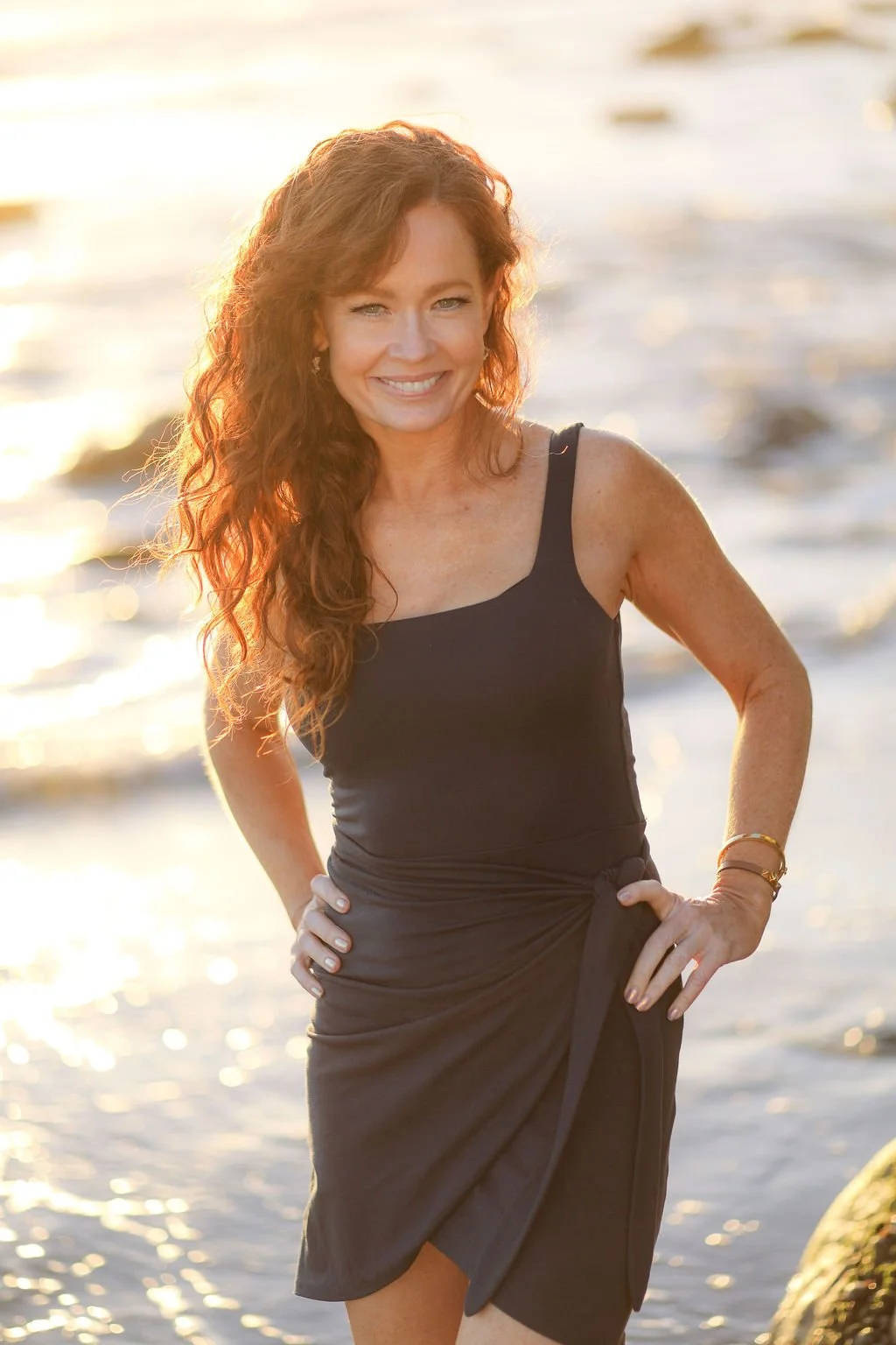 Woman with long curly red hair smiling at the camera, standing on a beach at sunset.