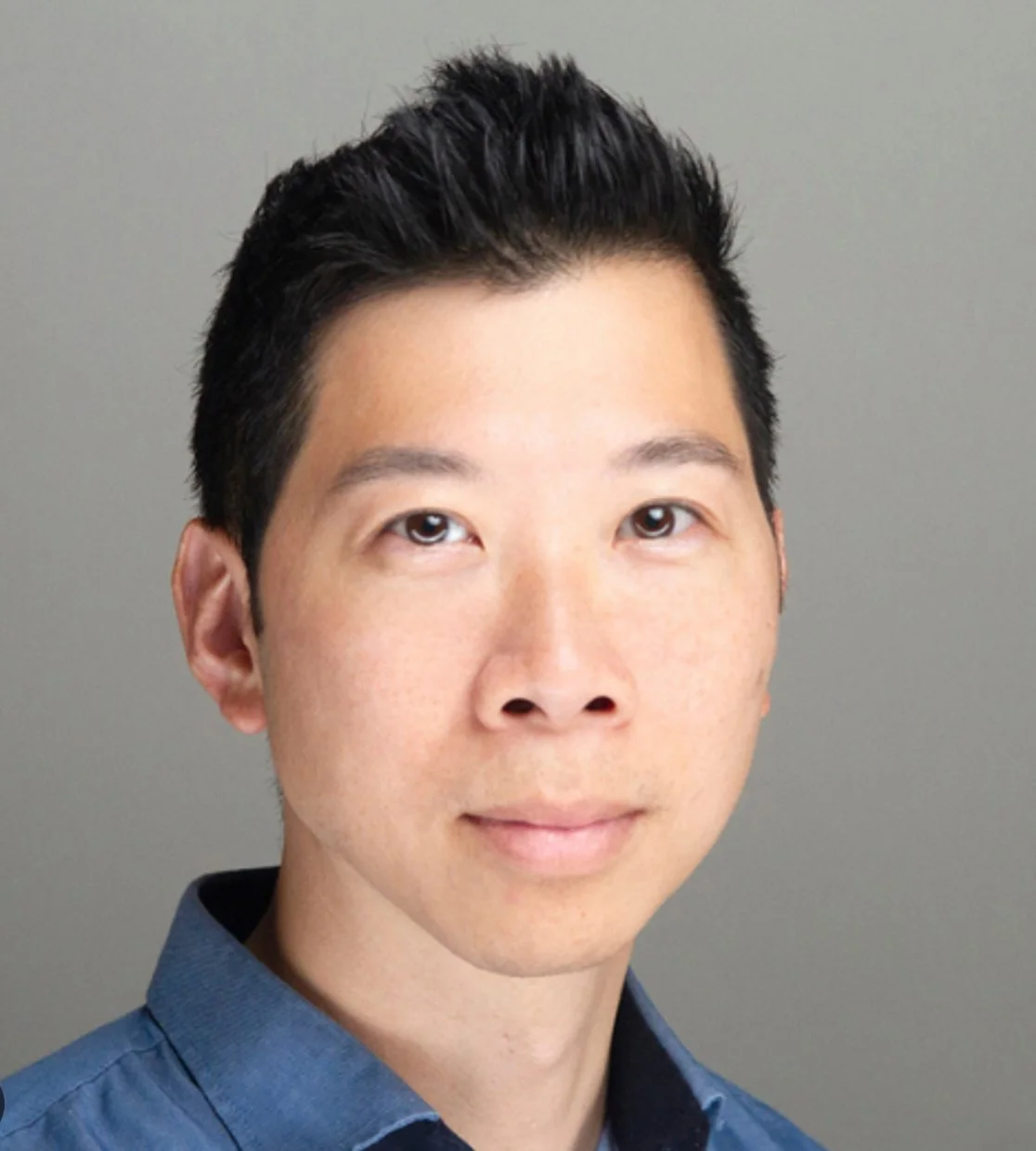 Headshot of a young Asian man with short black hair, wearing a blue collared shirt, smiling subtly against a plain gray background.