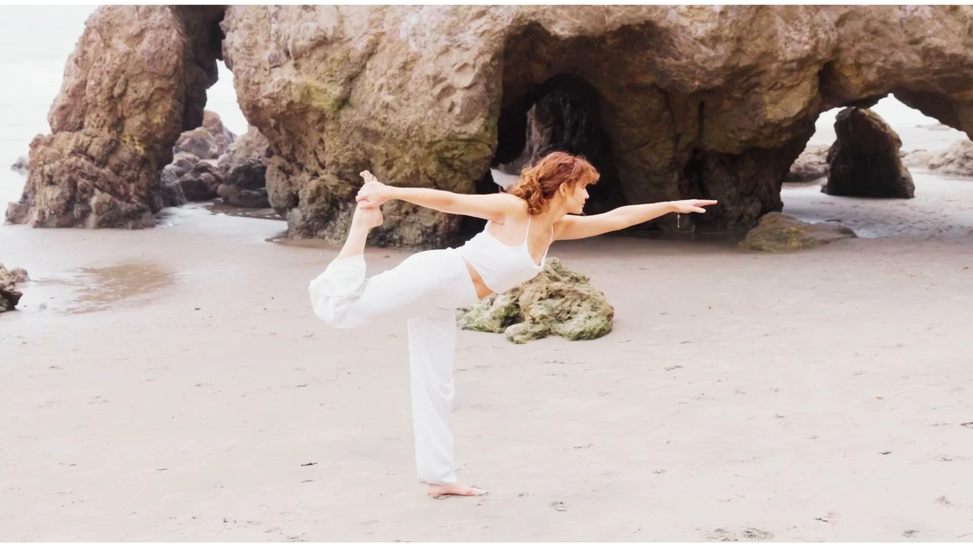 A woman practicing yoga on the beach, balancing in a yoga pose with one leg bent behind her and holding her foot with her hand, while extending her other arm forward. There are large rock formations and a sandy shoreline in the background.