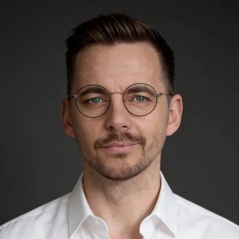 Ein Mann mit Brille und kurzen, dunklen Haaren trägt ein weißes Hemd vor einem dunklen Hintergrund.