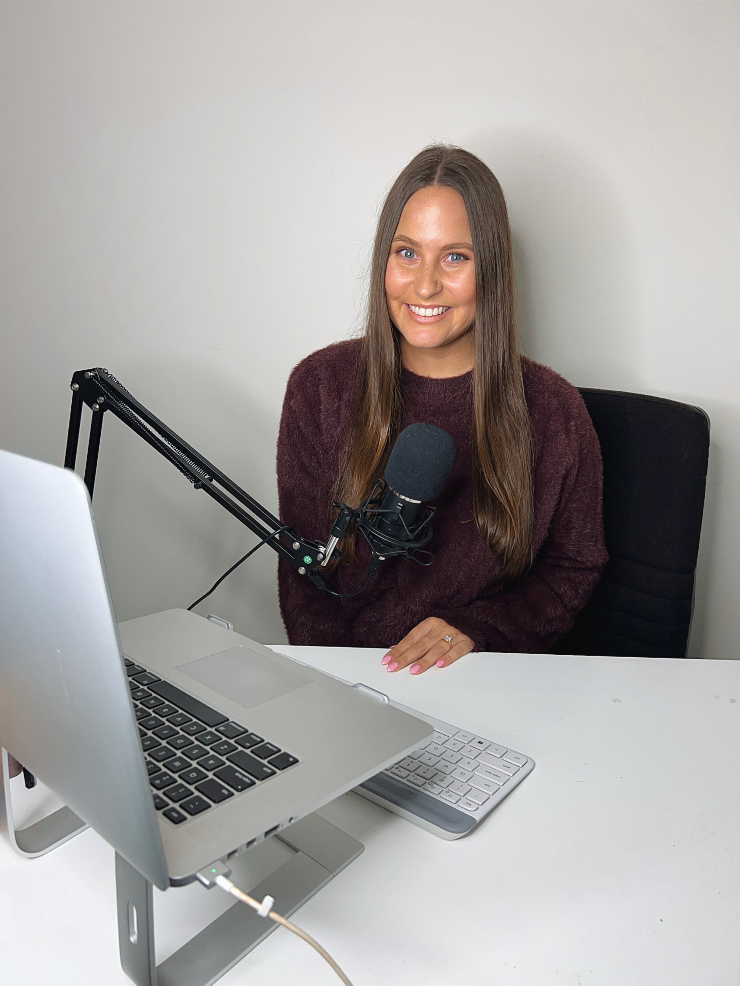 A woman with long brown hair, wearing a maroon sweater, smiling while sitting in front of a microphone and a laptop on a desk.