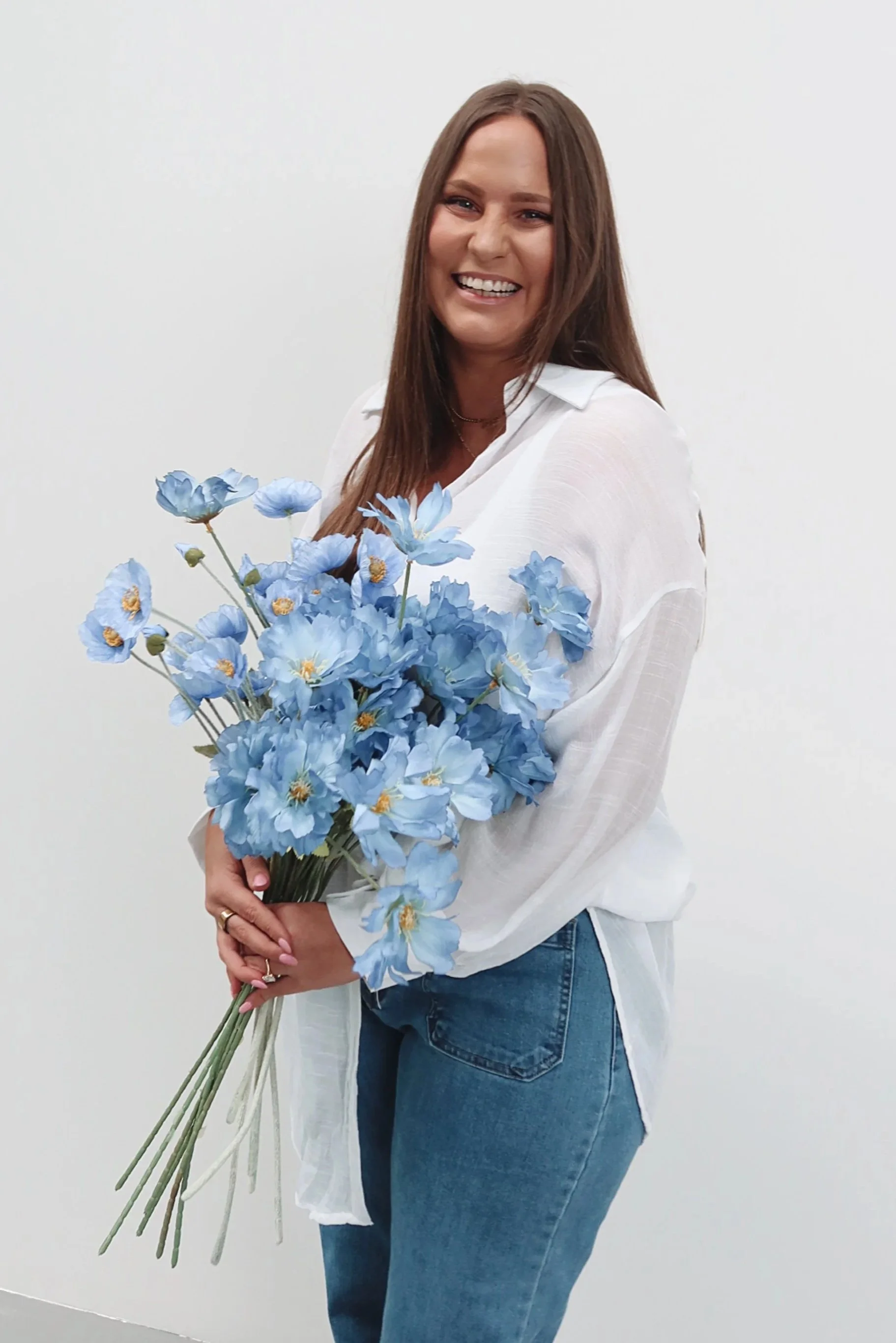 A woman smiling, holding a large bouquet of blue flowers, standing against a plain white background.