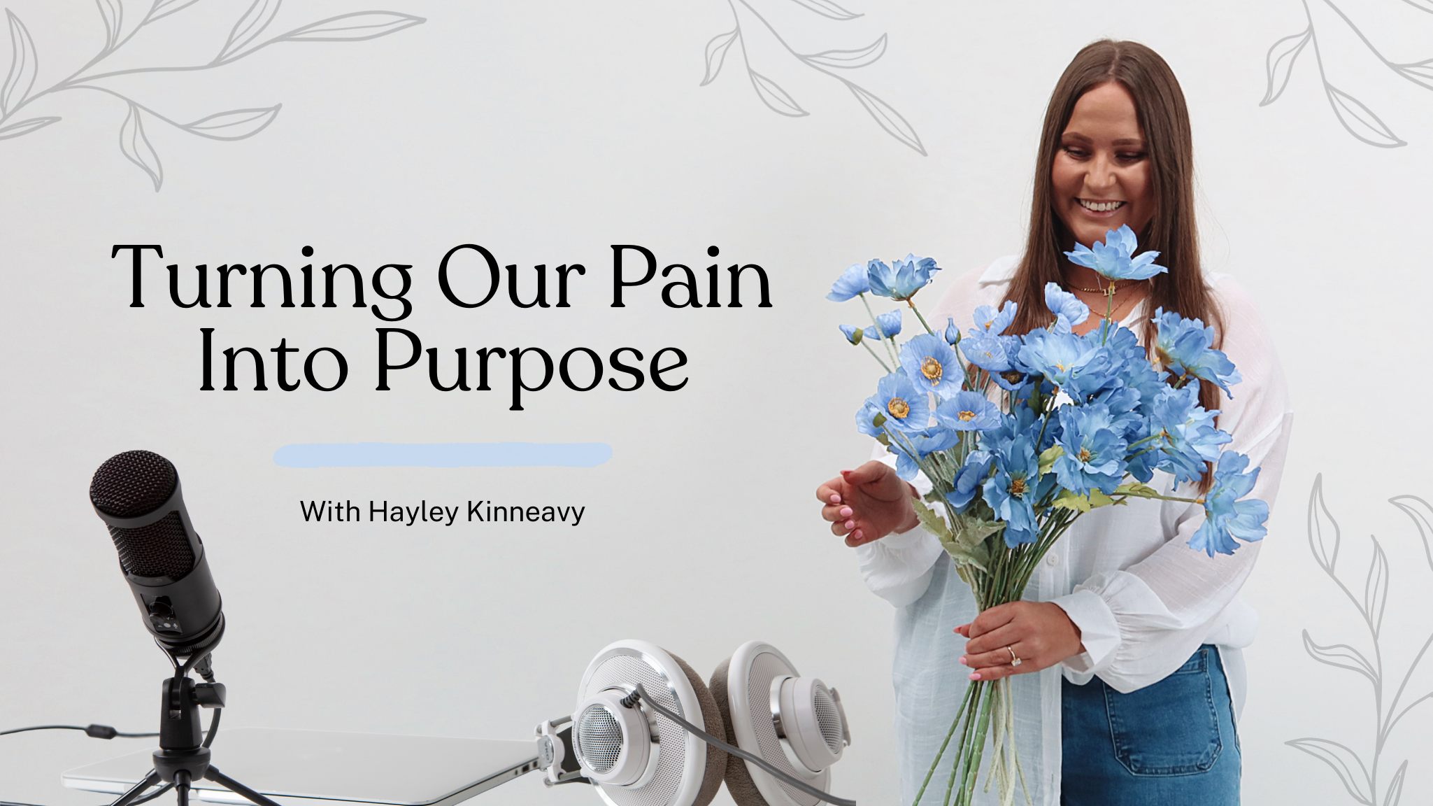 A woman with long brown hair, wearing a white shirt and jeans, smiling and holding a large bouquet of blue flowers, standing in front of a white wall with a podcast title "Turning Our Pain Into Purpose with Hayley Kinneavy." On the table in front of her are a microphone, a laptop, and a pair of headphones.
