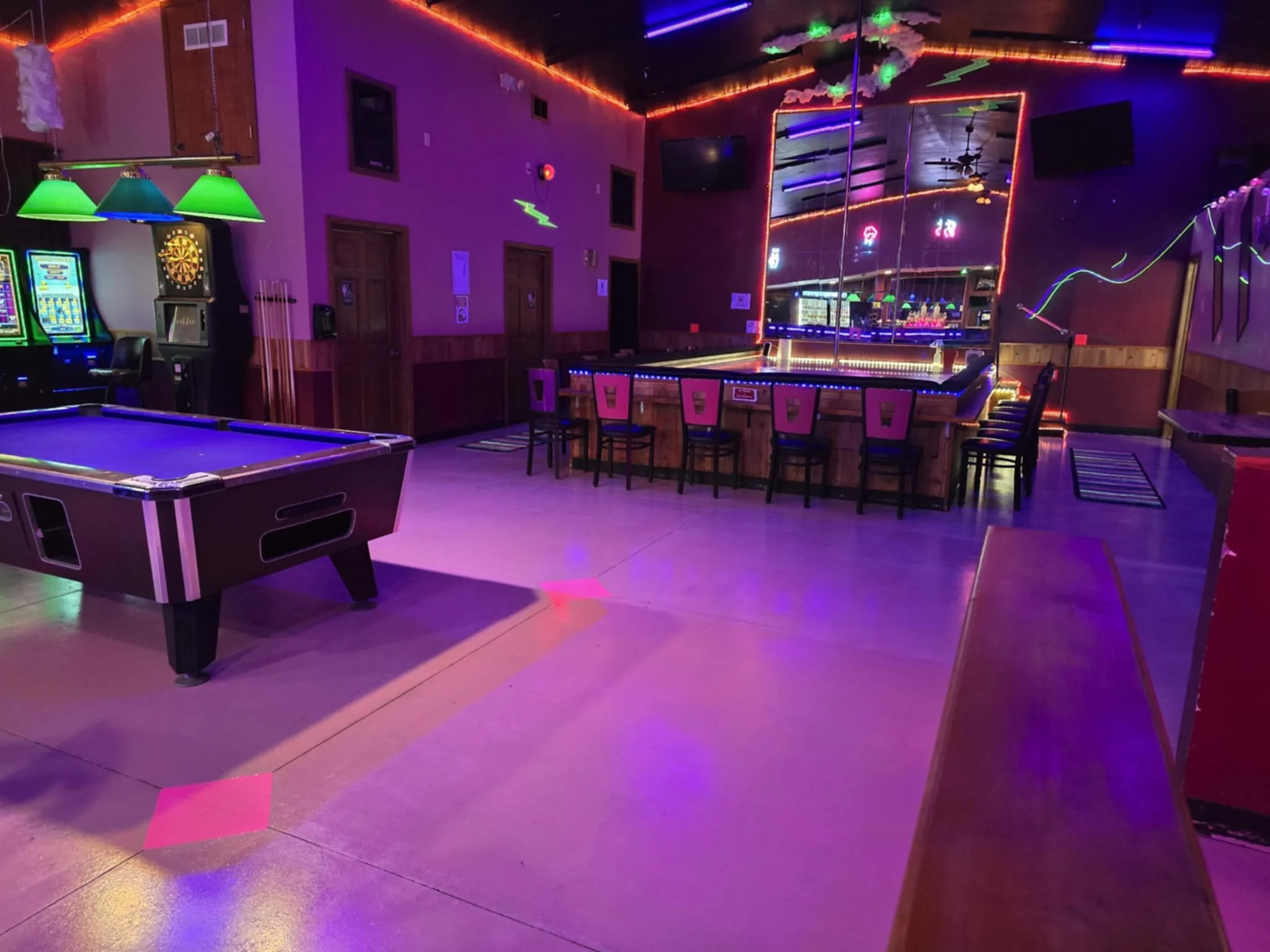 Empty game room with neon lighting, includes a pool table, arcade machines, bar area with chairs, neon signs, and reflective mirrors on walls.