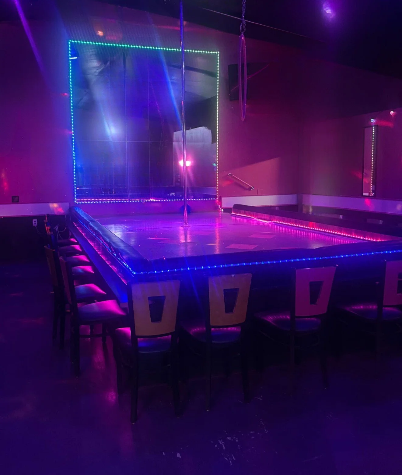 An empty dance club or performance venue with a stage featuring a pole, surrounded by illuminated LED lights in purple and blue. Chairs are lined up around the stage, and there are mirrors and gymnastic rings hanging from the ceiling.