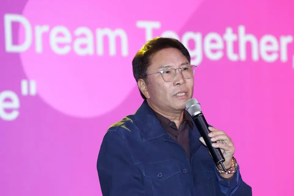 A man with glasses and dark hair is speaking into a microphone at a presentation or event. The background features pink and purple with the partially visible words 'Dream Together'.