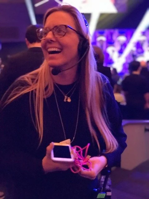 A woman wearing glasses and a headset with a microphone, smiling while holding a smartphone and pink earphones in a crowded indoor event space with purple lighting.