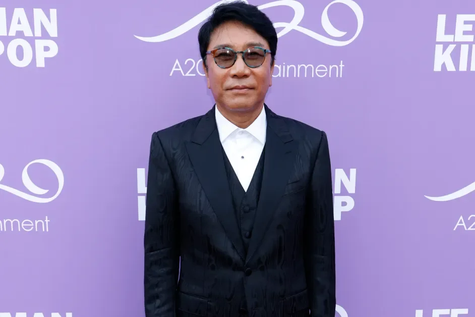 A man wearing a black suit, white shirt, and glasses standing in front of a purple backdrop with white text and logos, including the words 'LEON KID' and 'A2 Entertainment.'