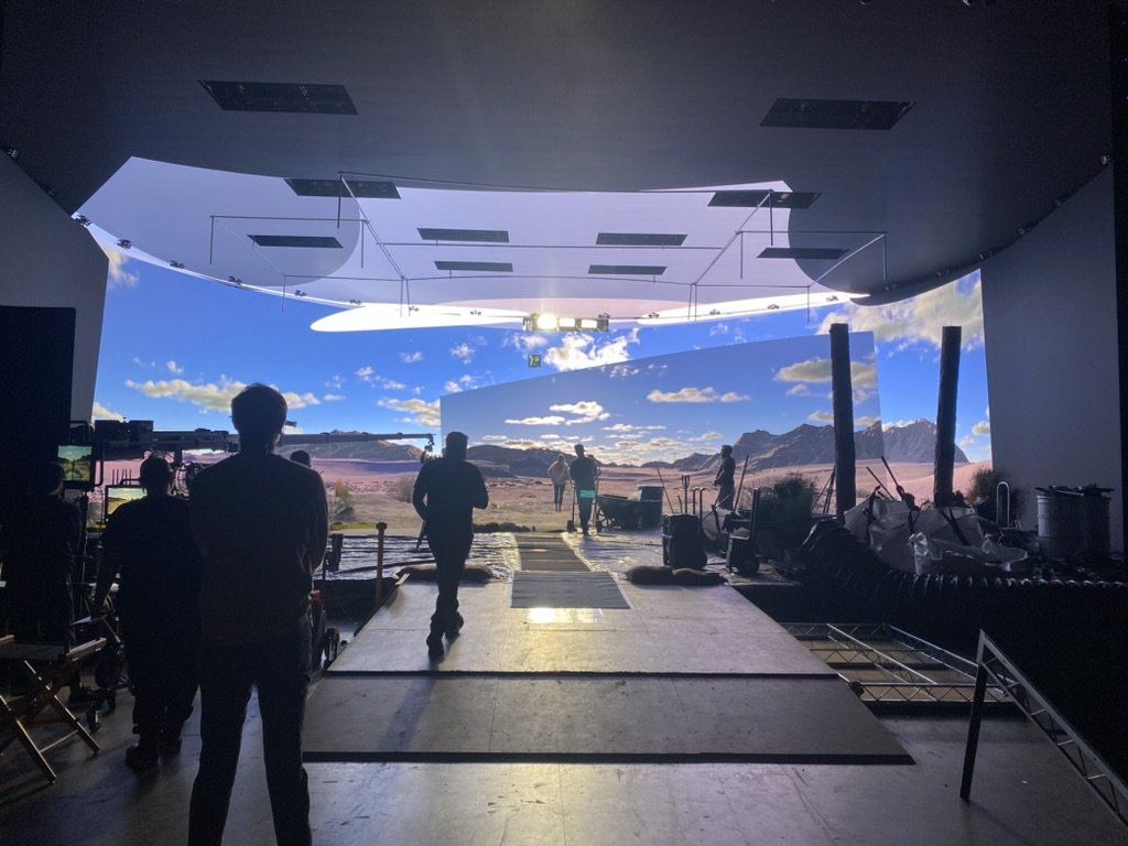 Behind-the-scenes view of a film or TV production set with people working, set design resembling a landscape with mountains and skies projected onto a curved screen, and studio equipment around.