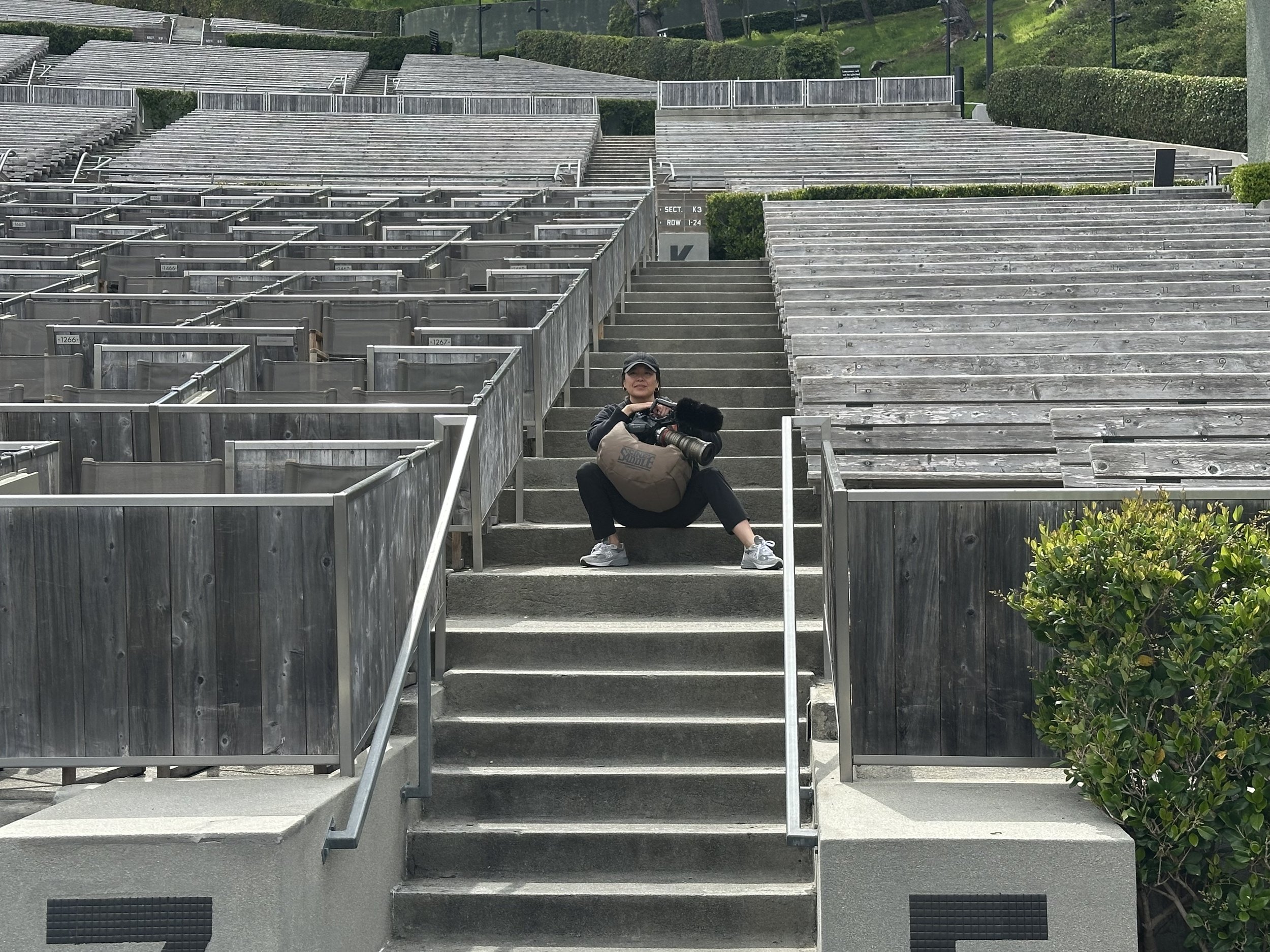 A woman sitting on outdoor gray concrete steps, holding a camera, with rows of empty outdoor amphitheater seating made of weathered wood in the background, and green bushes on the right side.