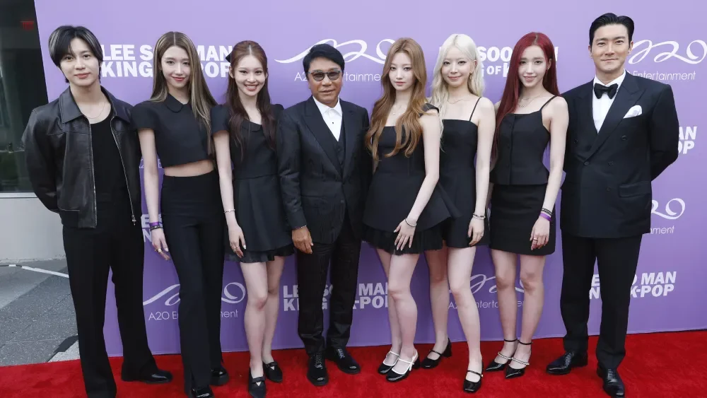 A group of nine people, six women and three men, dressed in black formal and semi-formal clothing, standing on a red carpet in front of a purple backdrop with white writing and logos at an event.