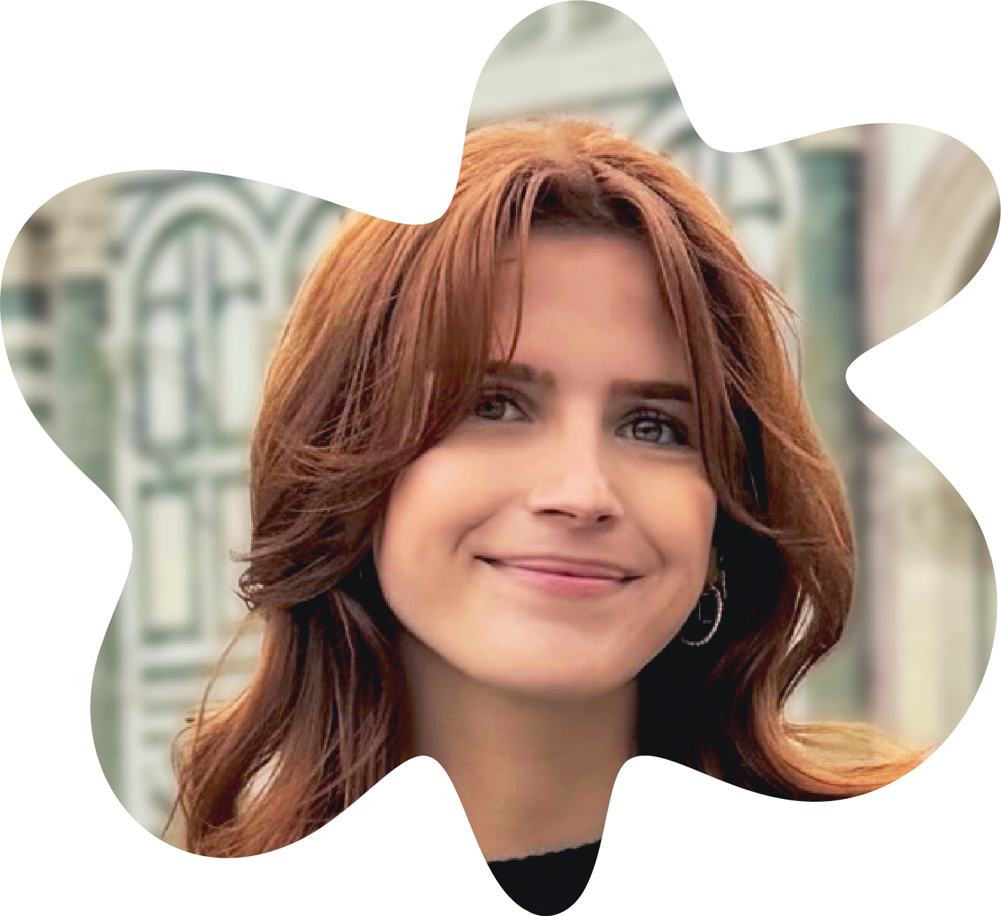 Ashley Toombs-Heavisides is smiling with closed lips. She has red hair and silver hoop earrings. behind her is out of focus European architecture. The photo is cropped into the loose shape of a flower.